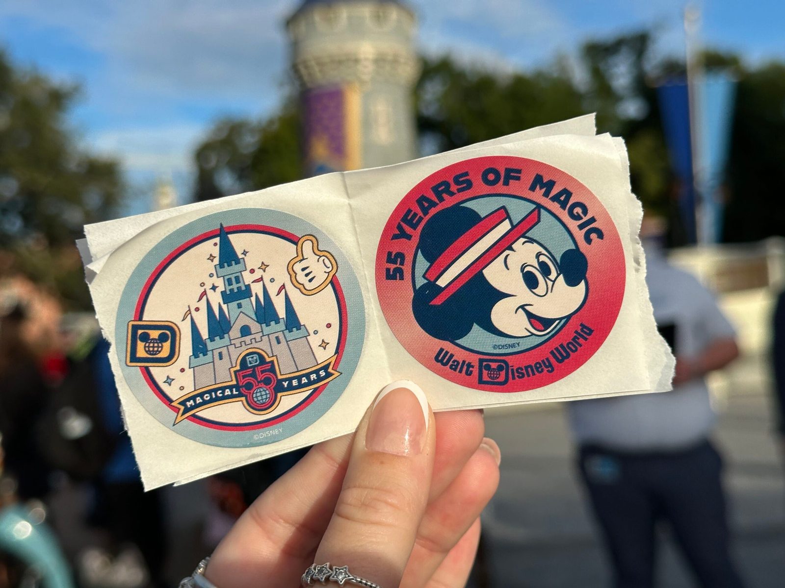 A hand holds two round stickers in front of a stone tower at Magic Kingdom: a pastel Cinderella Castle with a Magical 55 Years banner and a thumbs-up Mickey glove, and a red sticker reading 55 Years of Magic with Mickey in a striped hat