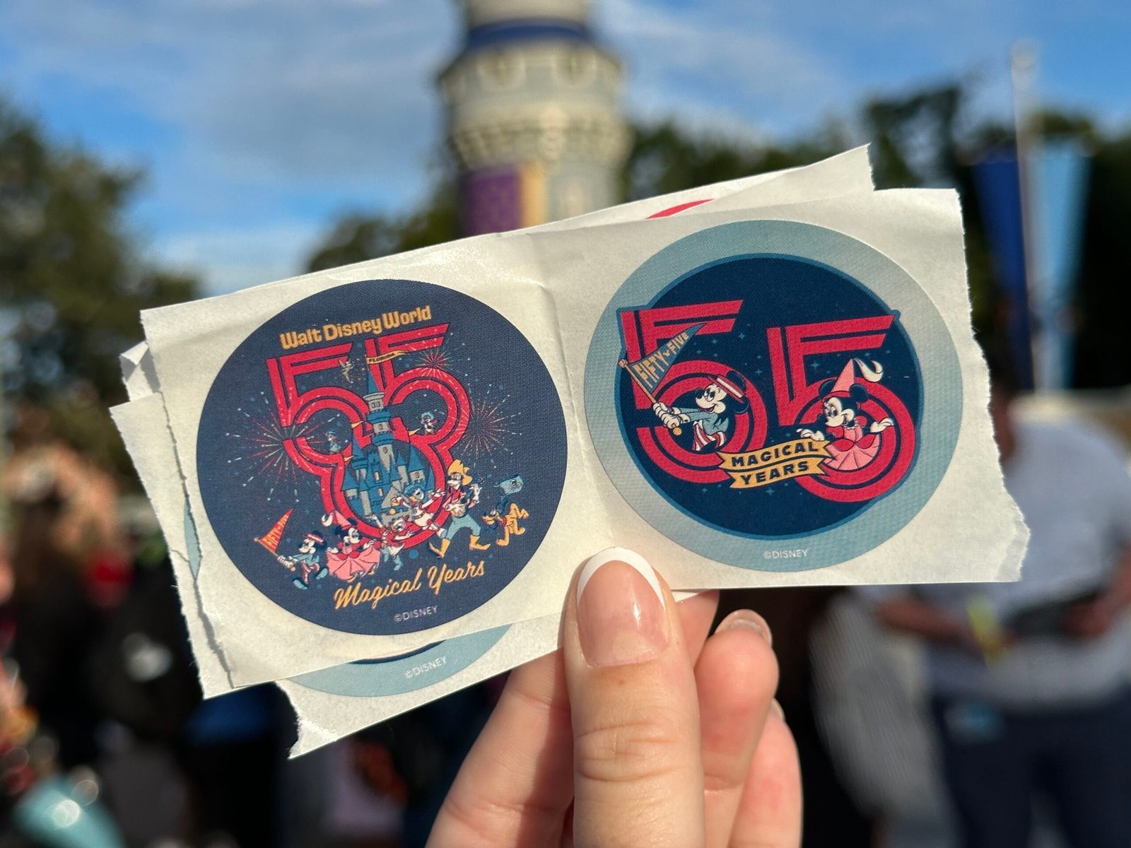 Two navy stickers held up at Magic Kingdom: one with Mickey, Minnie, Donald, Daisy, Goofy and Pluto parading past the castle inside a red 55 with Magical Years script, the other with Mickey and Minnie in a big red 55 over a Magical Years banner