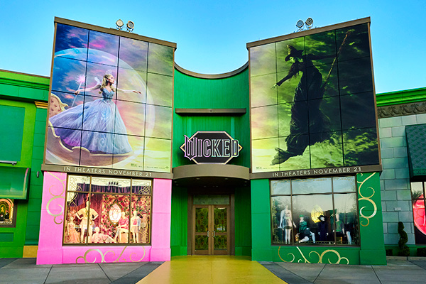 Green and pink storefront with a Wicked sign over the doors, large Glinda and Elphaba panels with In Theaters November 21 banners, and costume display windows below