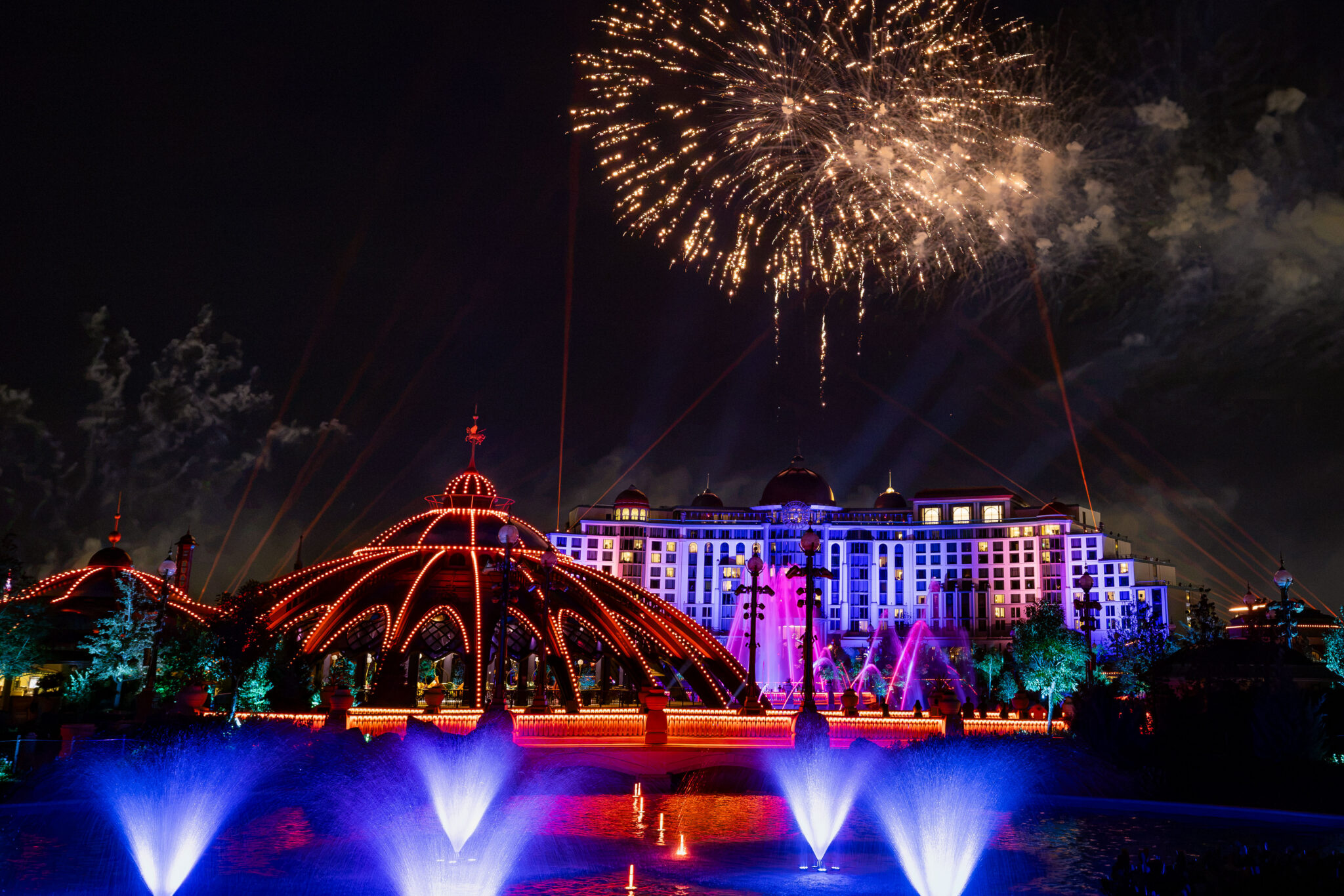 Gold fireworks burst over a brightly lit hotel and a domed pavilion outlined in red lights, with blue-lit fountains in a lagoon in the foreground