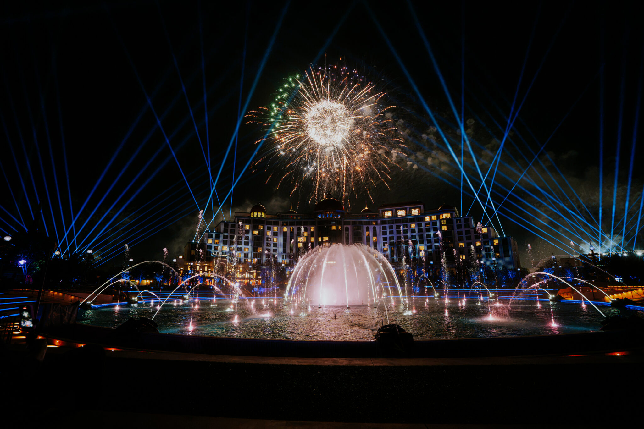 A firework bursts above a large hotel while blue searchlight beams fan across the sky and arcs of fountain water rise from a round pool