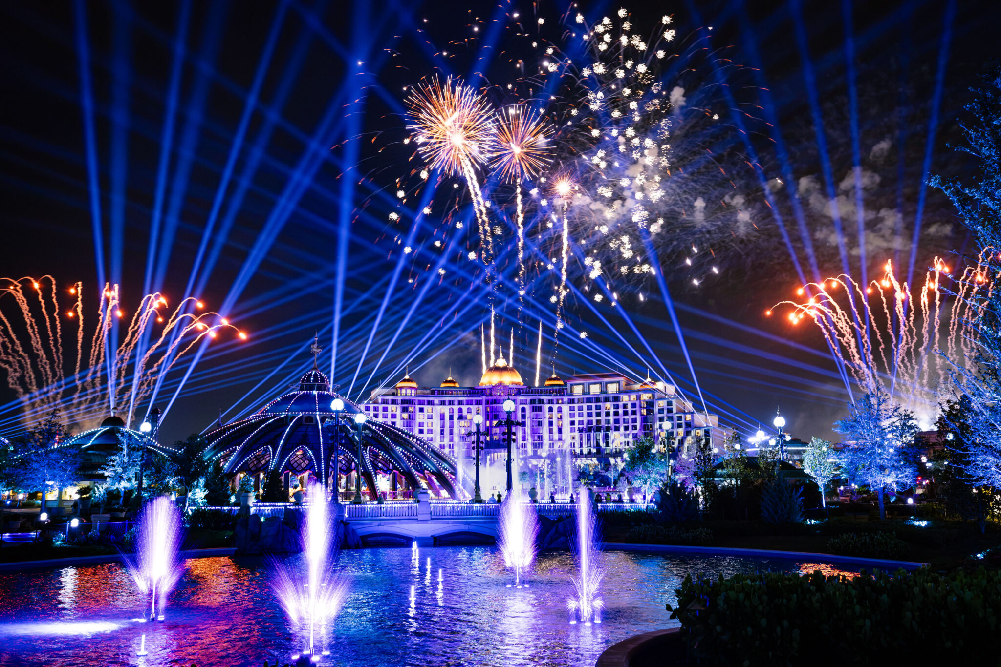 Blue searchlight beams and fireworks fill the sky above a hotel, with fountain jets rising from a lagoon in front of a lit domed pavilion