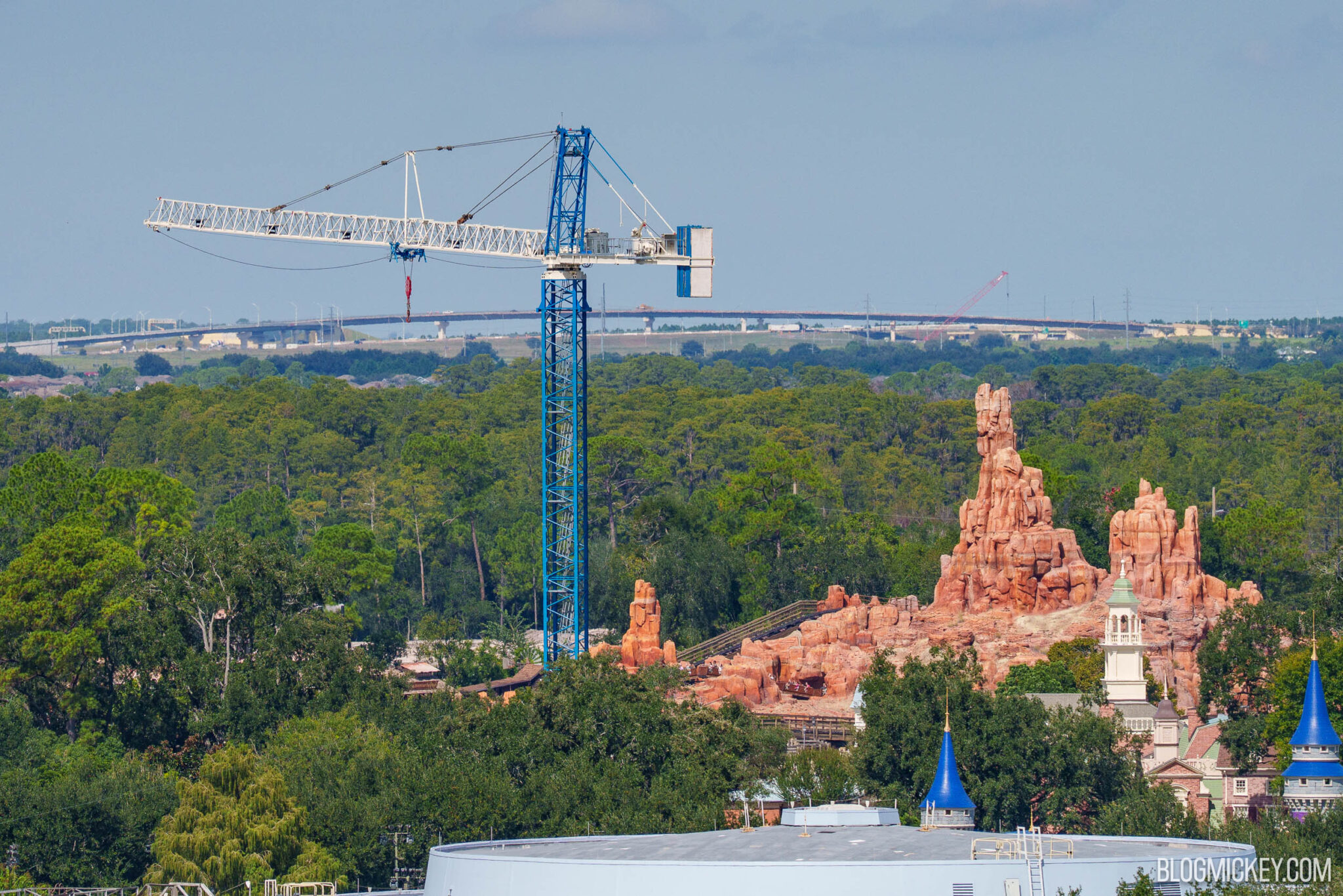 A blue tower crane with a white jib rises above trees behind the red rock spires of Big Thunder Mountain Railroad, with blue turret tops in the foreground