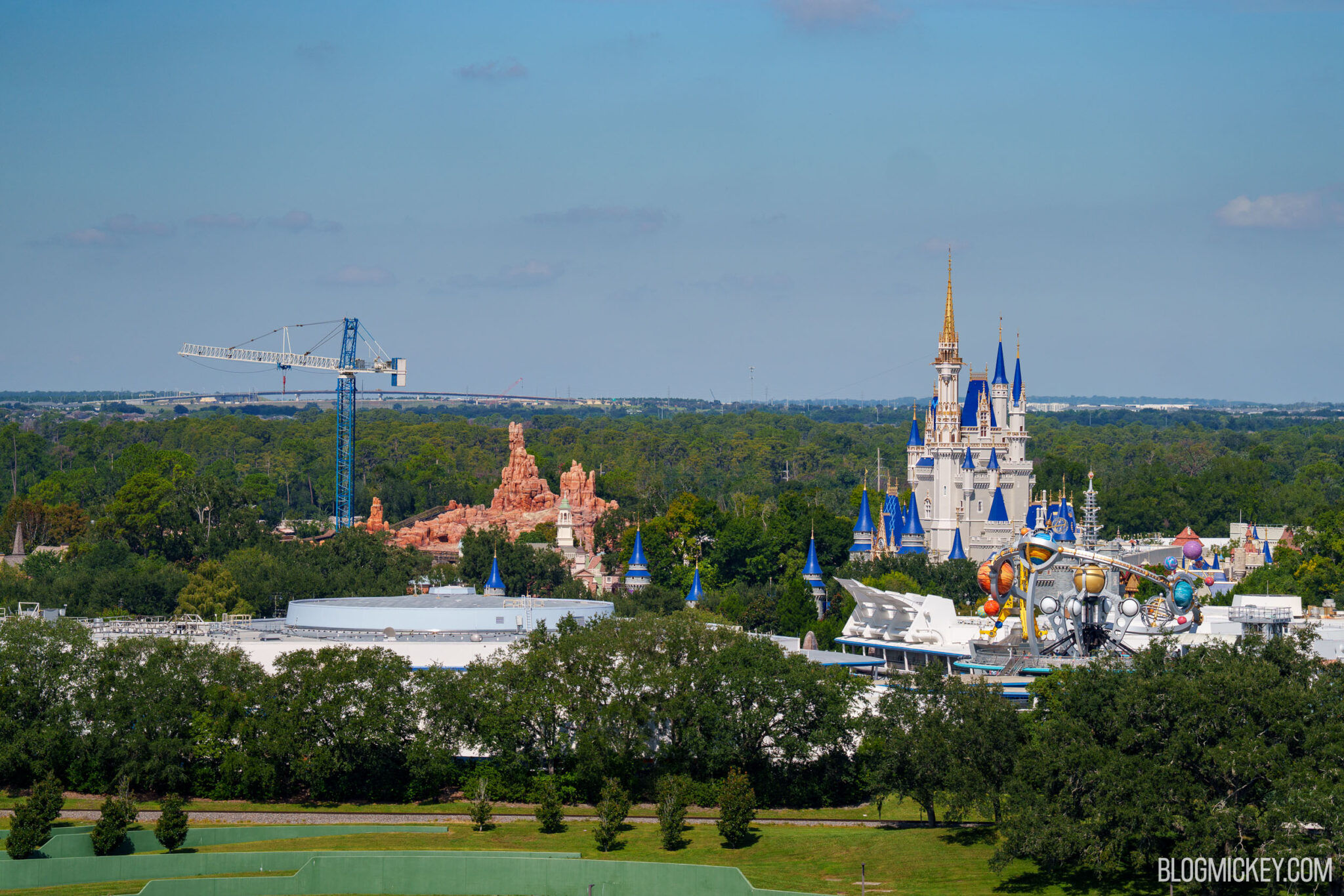 Wide elevated view across Magic Kingdom showing a blue tower crane beside the red rock of Big Thunder Mountain Railroad on the left and Cinderella Castle and the Astro Orbiter in Tomorrowland on the right