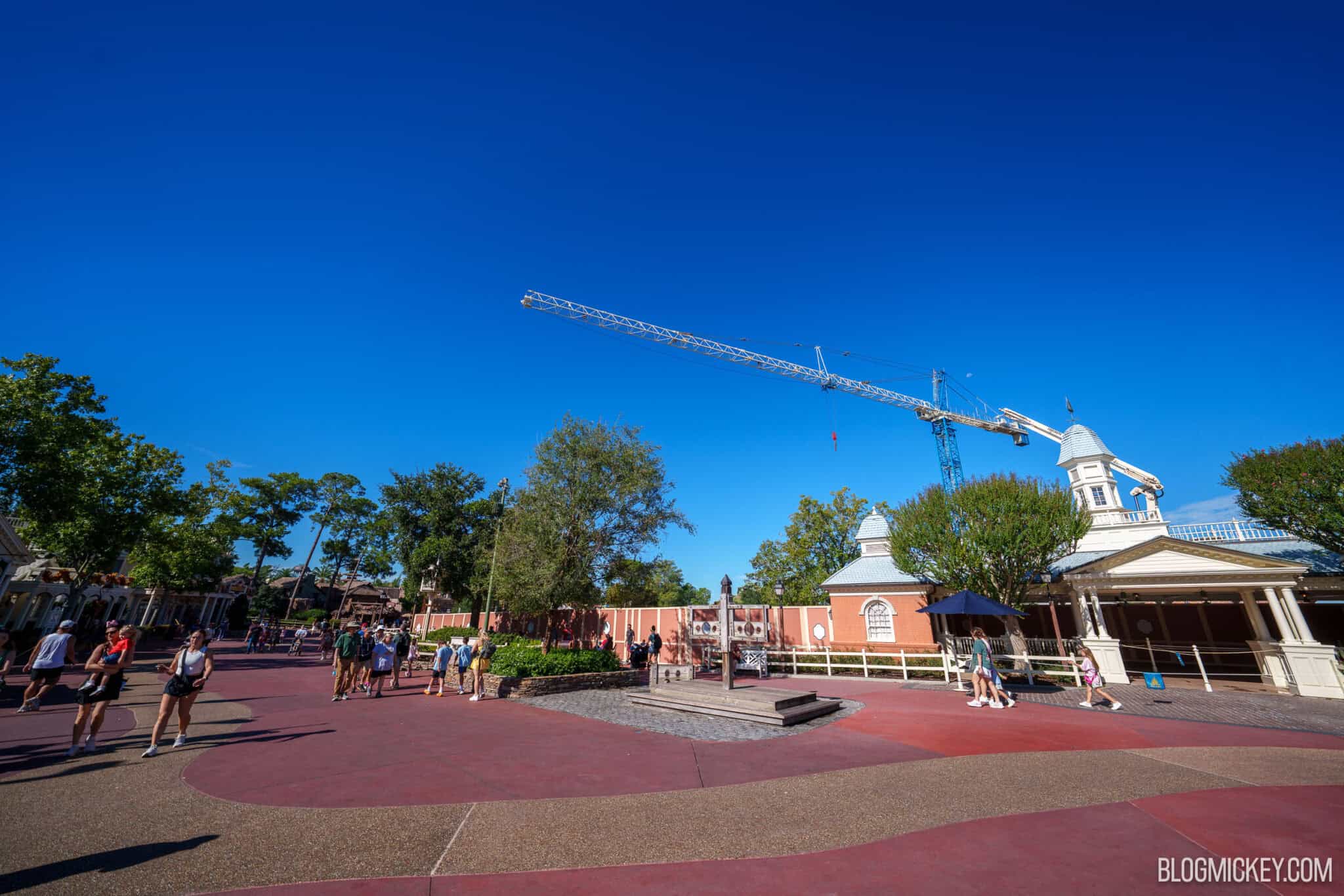A blue tower crane mast rises behind the former Liberty Square Riverboat landing, its long white lattice jib stretching left across a clear blue sky over the construction walls, with guests walking across the red pavement of Liberty Square