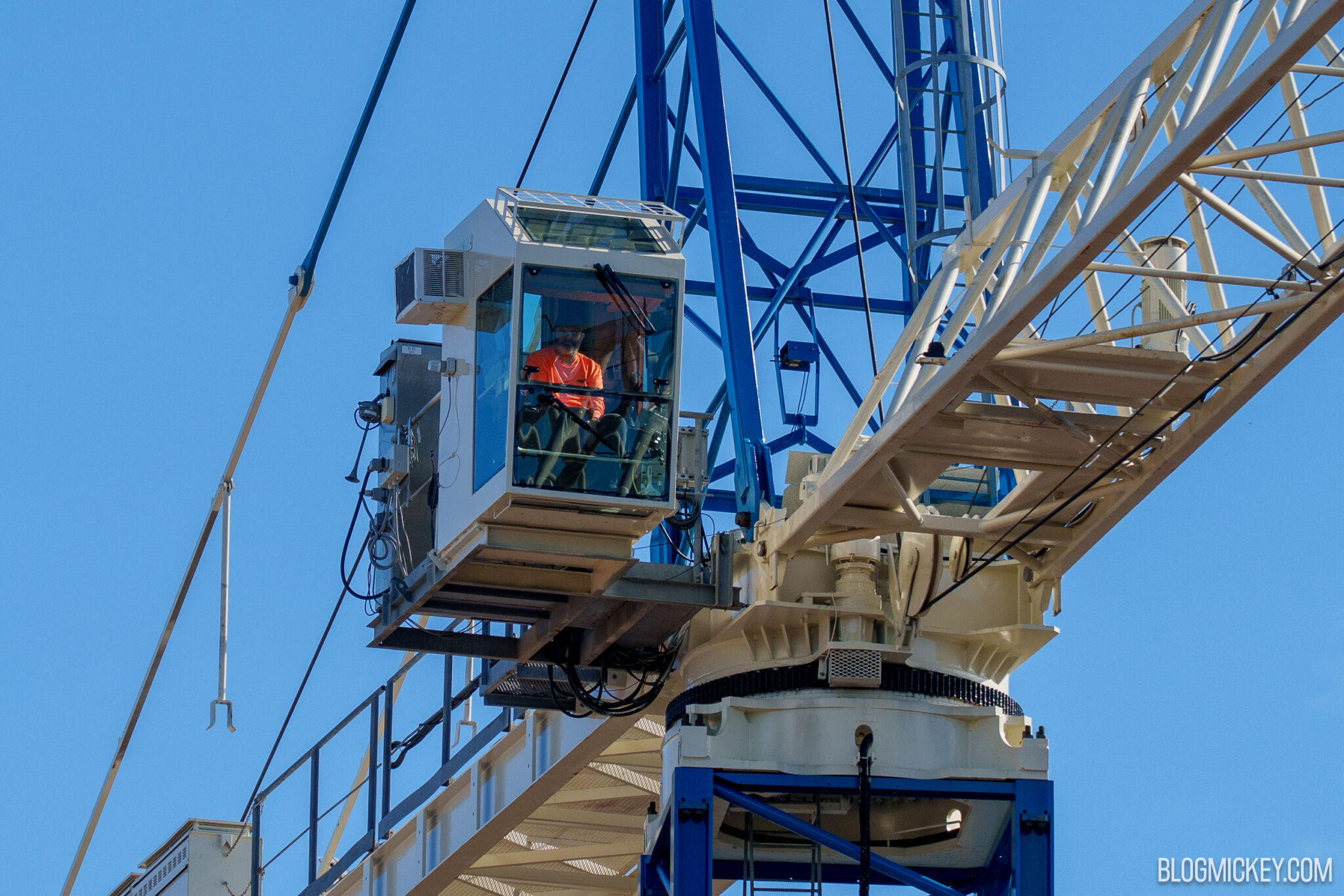 Close-up of a white crane operator cabin at the top of a blue steel mast, with a worker in an orange shirt seated inside behind the glass