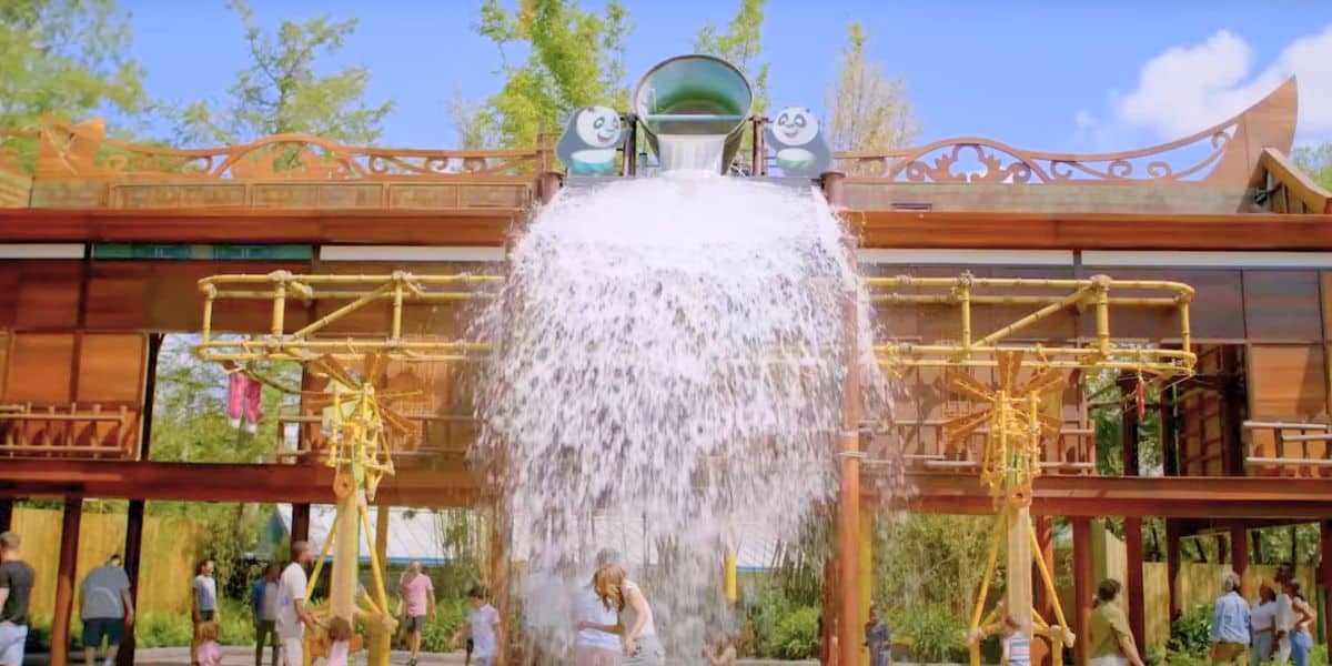 A large bucket tips a sheet of water down from a wooden Kung Fu Panda themed play structure with yellow bamboo poles and two panda faces on top, as guests stand underneath