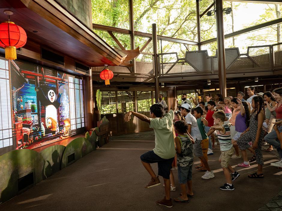 A group of children copy kung fu poses in front of a large screen showing an animated Po, inside an open-air pavilion hung with red paper lanterns