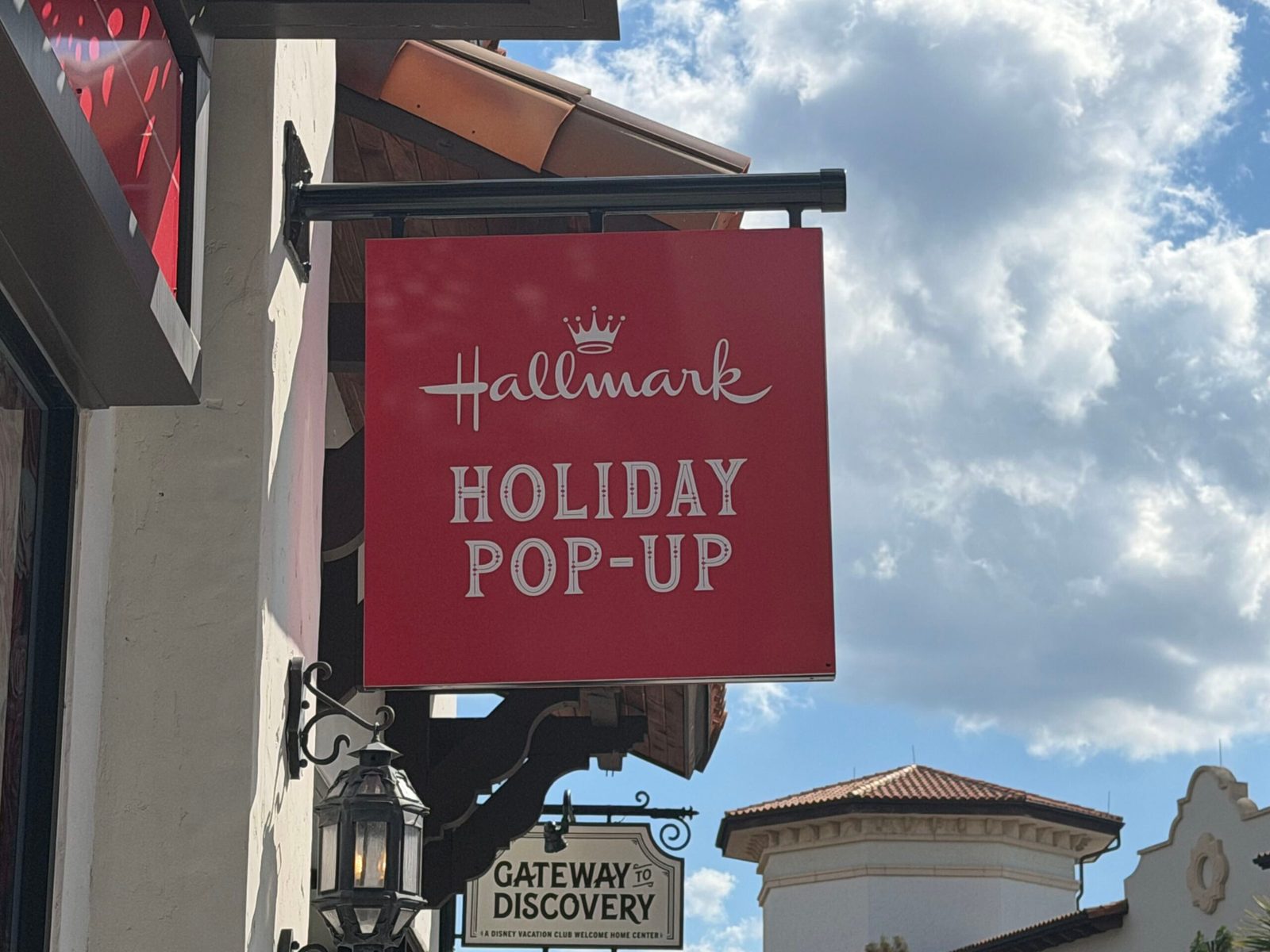 A red hanging sign reading Hallmark Holiday Pop-Up with the Hallmark crown logo, mounted on a Disney Springs building, with a separate Gateway to Discovery Disney Vacation Club sign hanging lower in the background