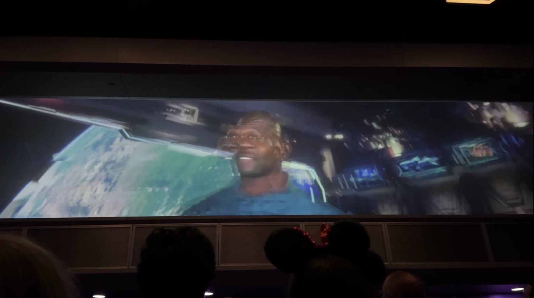 A wide preshow screen showing Terry Crews as Centurion Tal Marik in a blue uniform in front of a view of Earth, with his face doubled and smeared by a digital glitch effect, and silhouettes of guests including Minnie ears in the foreground