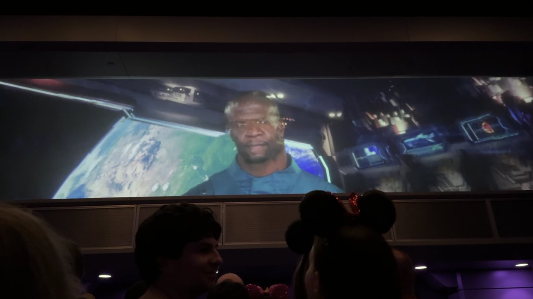Terry Crews as Centurion Tal Marik on the wide preshow screen with Earth and a spaceship control panel behind him, slightly blurred, with guests watching from the darkened room below