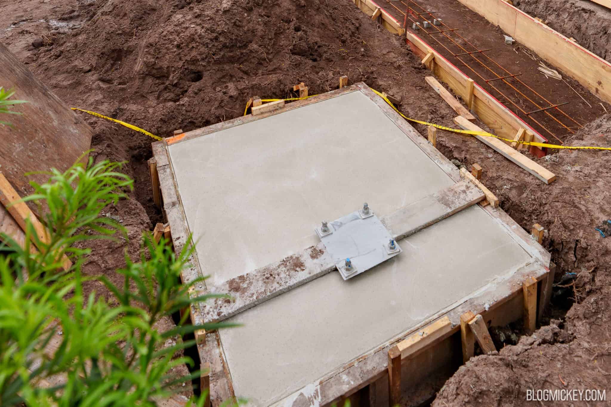 A freshly poured square concrete pad inside a wooden form, with a square steel plate and four anchor bolts set into the center, surrounded by dug soil and yellow caution tape, with a rebar-lined trench behind it