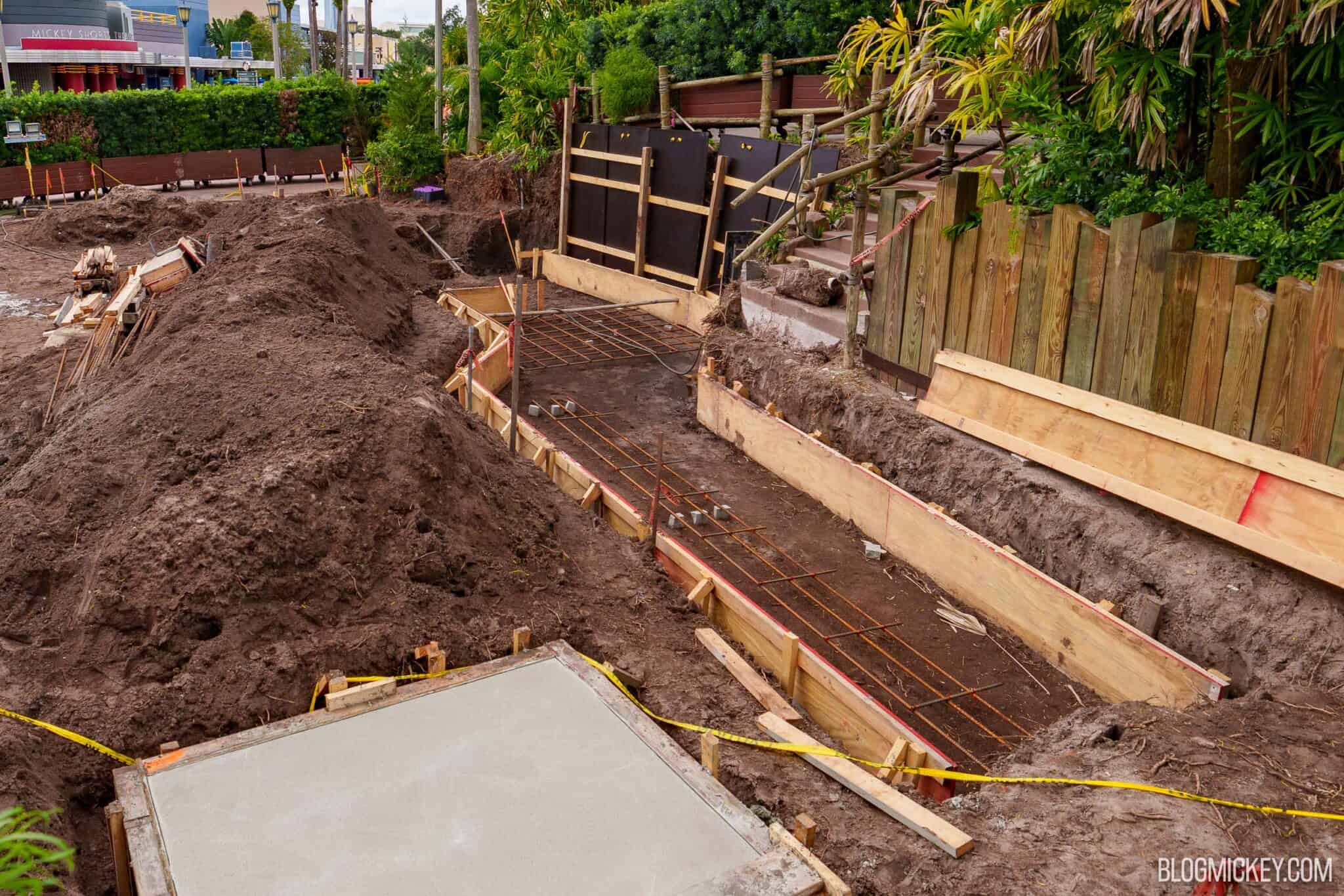 A long trench lined with wooden forms and rebar running beside a wooden fence and steps at Hollywood Studios, with black forming panels behind it, mounds of dirt to the left, and the corner of the concrete pad in the foreground