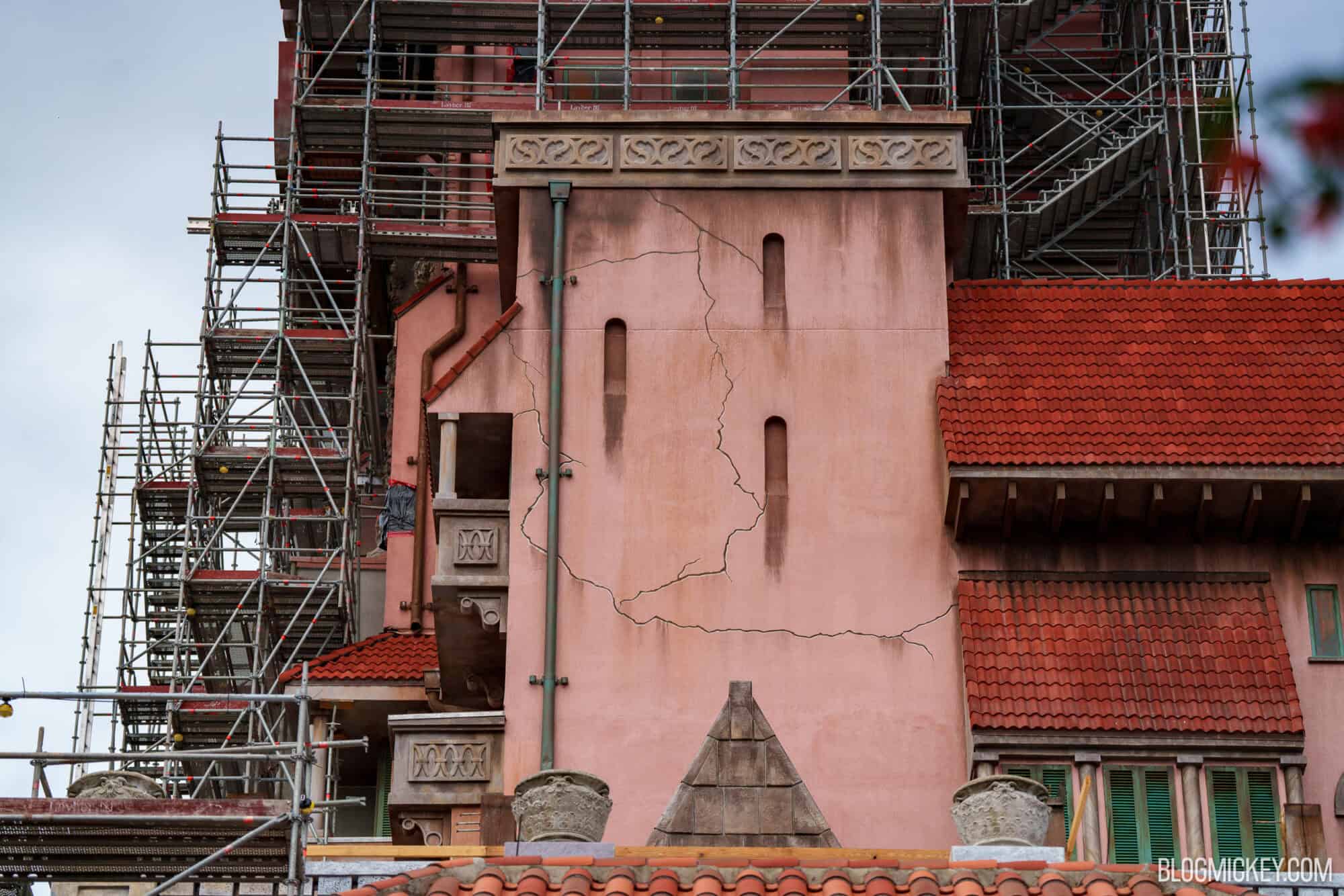 Close view of a pink stucco wall on the Hollywood Tower Hotel with long painted cracks, dark vertical streaks below narrow arched windows, a teal downspout and red tile roofs, with scaffolding at left and above