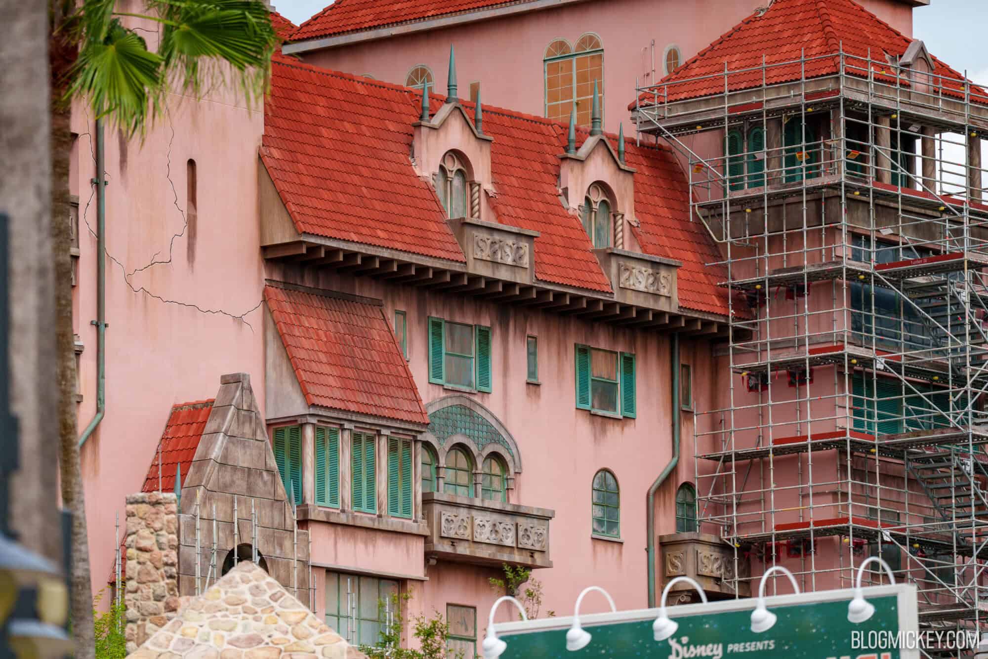 A pink stucco wing of the Hollywood Tower Hotel with red tile roofs, green spires, teal shutters, painted cracks and dark streaks, with scaffolding still standing on a corner section at right
