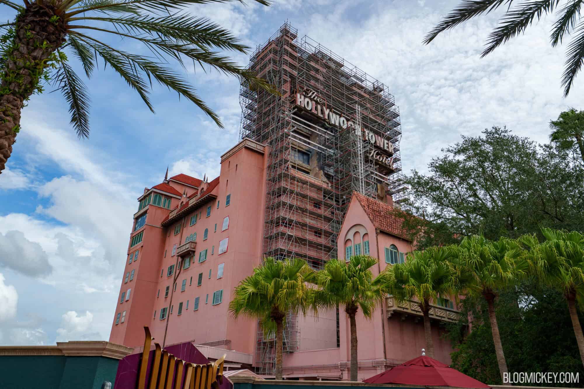 Looking up at the Hollywood Tower Hotel with scaffolding wrapping the upper tower around the Hollywood Tower Hotel sign, palm trees in the foreground and the pink lower building at left