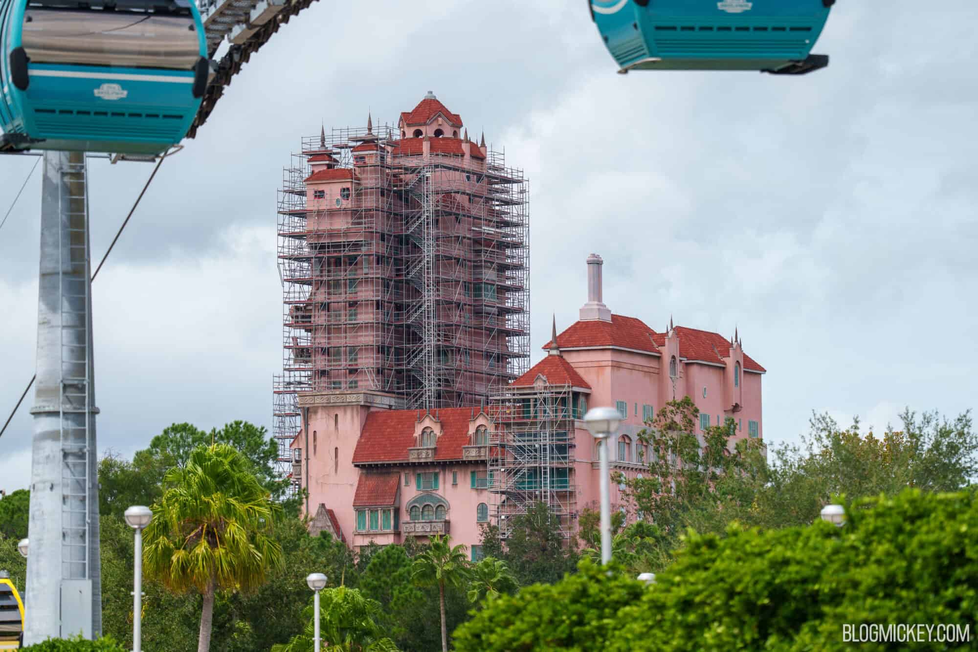 Distant view of the Hollywood Tower Hotel with scaffolding around the upper tower and one lower section, framed by two teal Disney Skyliner gondolas and trees