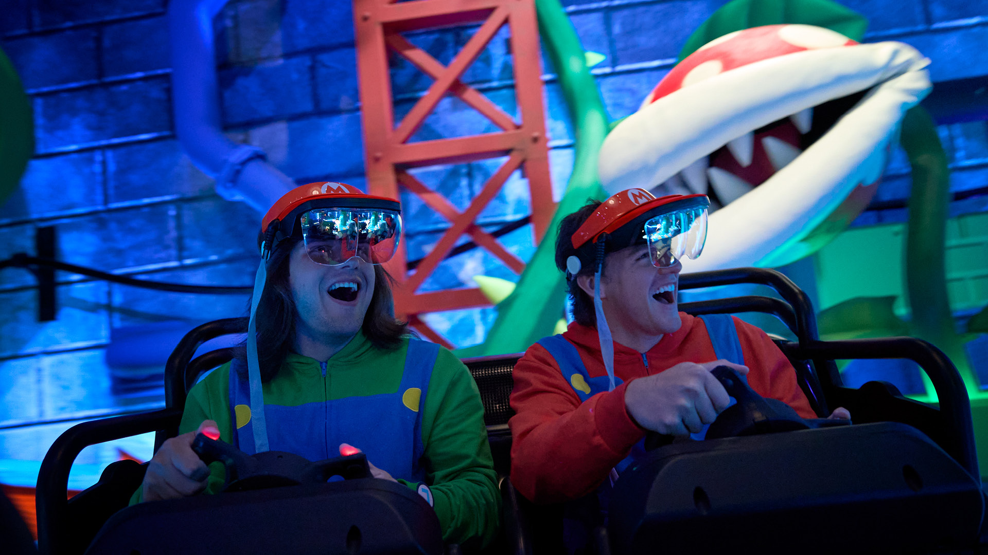 Two guests dressed as Luigi and Mario laughing while riding Mario Kart: Bowser's Challenge, wearing red Mario headsets with clear visors, with a giant Piranha Plant behind them
