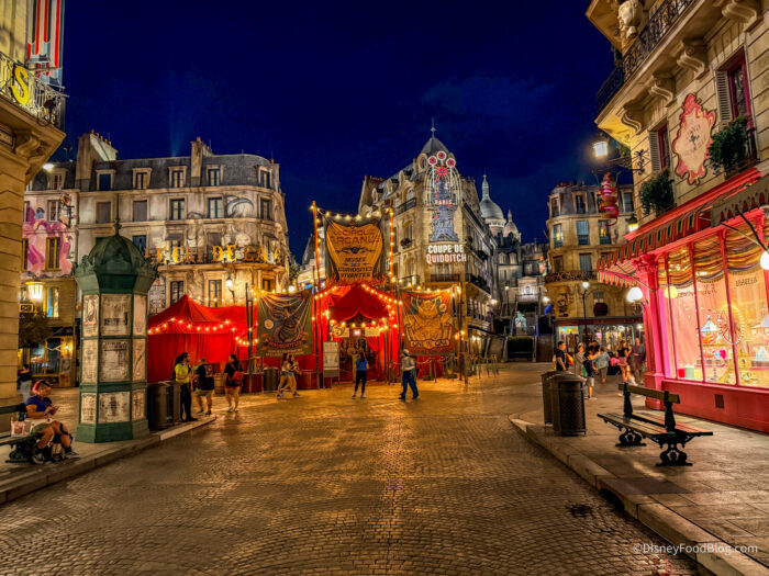 A cobblestone street in the Ministry of Magic at night with a red striped circus tent and posters for Le Cirque Arcanus, Parisian buildings lit up behind it, and only a handful of guests walking through