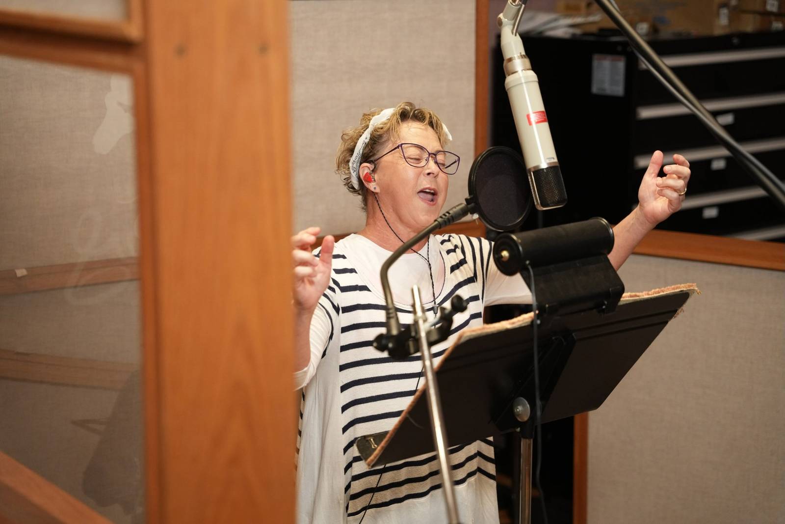 Debi Derryberry recording in a studio vocal booth, eyes closed and arms raised mid-line, in front of a large microphone with a pop filter and a music stand, seen past a wooden door frame