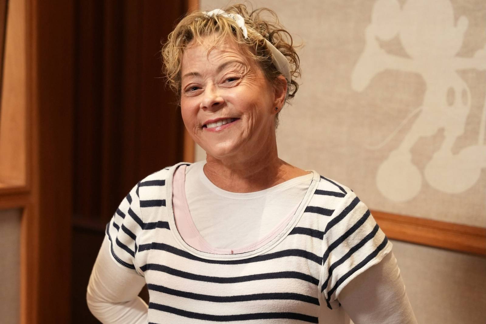 Debi Derryberry smiling at the camera in a striped top and white headband, standing in a recording studio with a large Mickey Mouse silhouette on the wall behind her