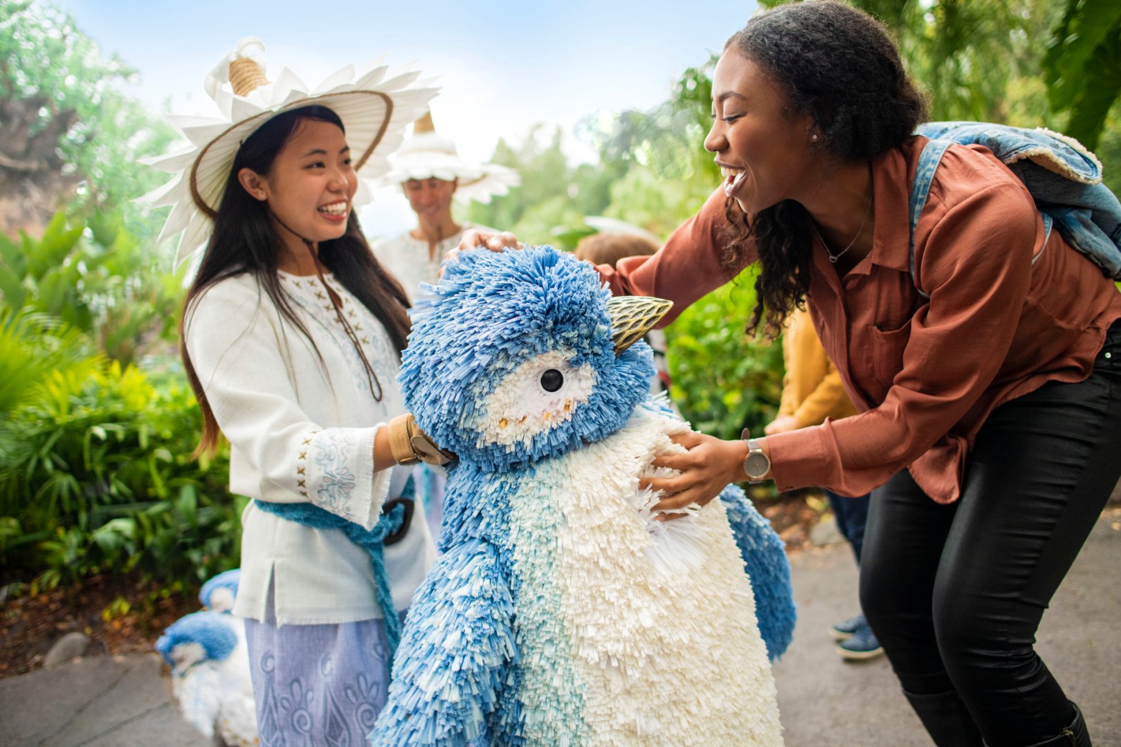 A smiling guest in a rust-colored shirt leans in to touch a large blue and white penguin puppet made of layered fringe, held by a performer in a white wide-brimmed hat, with a second performer and a smaller penguin puppet behind them among green plants
