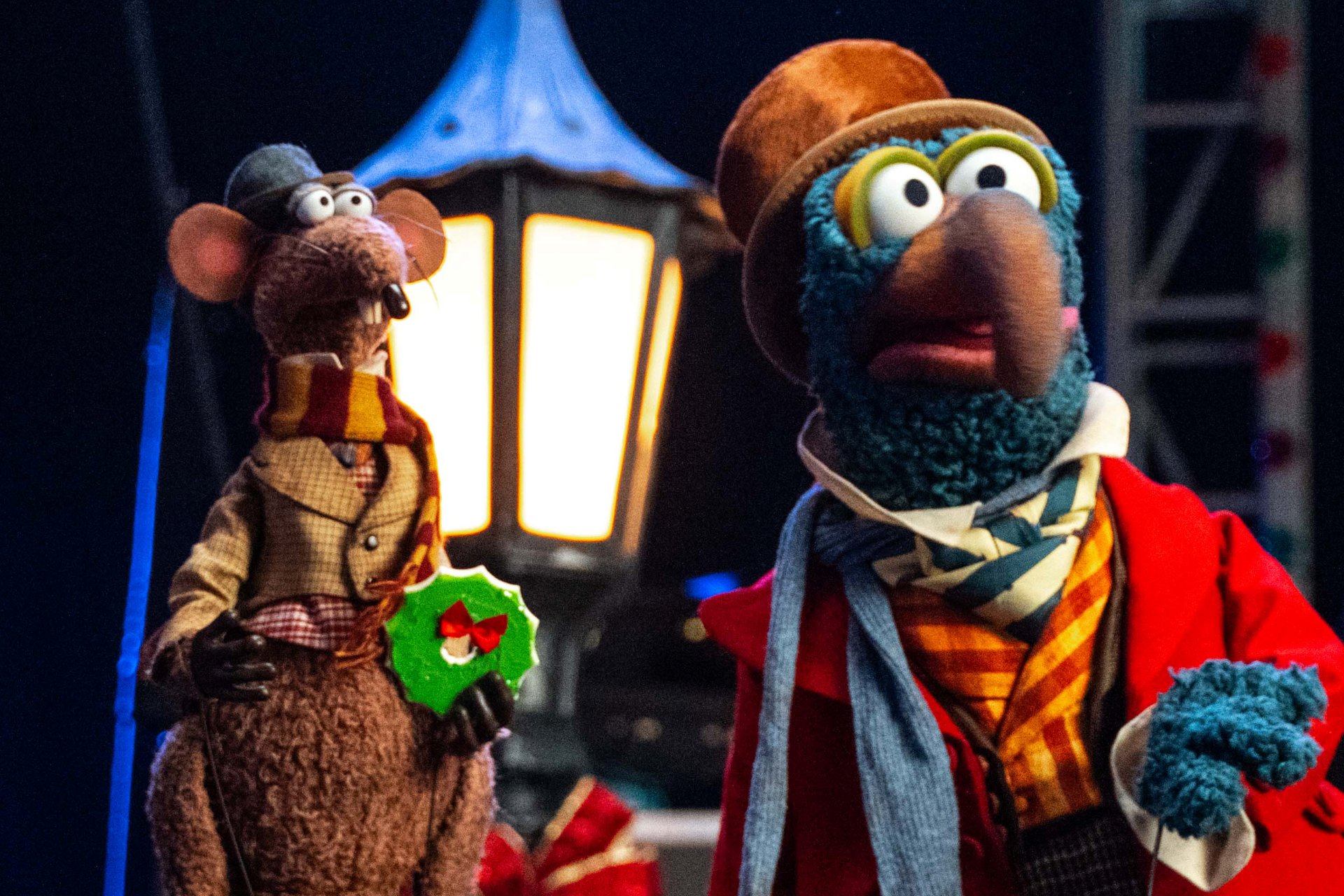 Rizzo the Rat holding a green wreath cookie and Gonzo in a brown bowler hat, red coat and striped scarf, standing in front of a glowing street lamp
