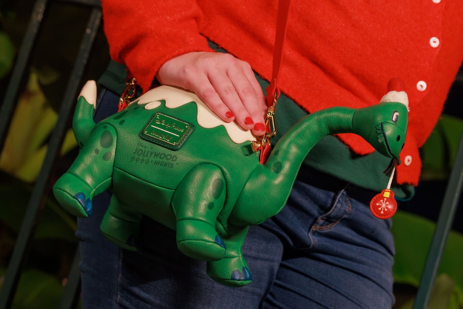 A green Gertie the Dinosaur Loungefly bag with white snow on its back, a small Santa hat and a red ornament hanging from its mouth, with Jollywood Nights printed on its side