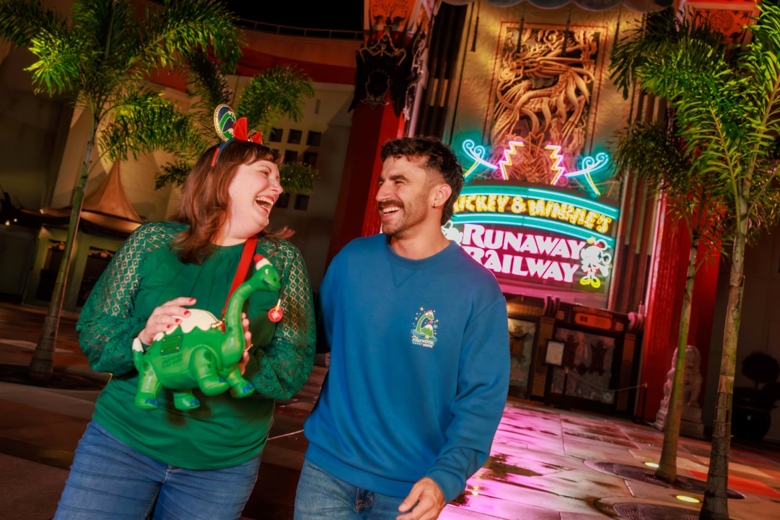 Two guests laughing at night outside Mickey and Minnie's Runaway Railway, one holding the green Gertie Loungefly and wearing the Jollywood Nights headband, the other in a blue Jollywood Nights pullover