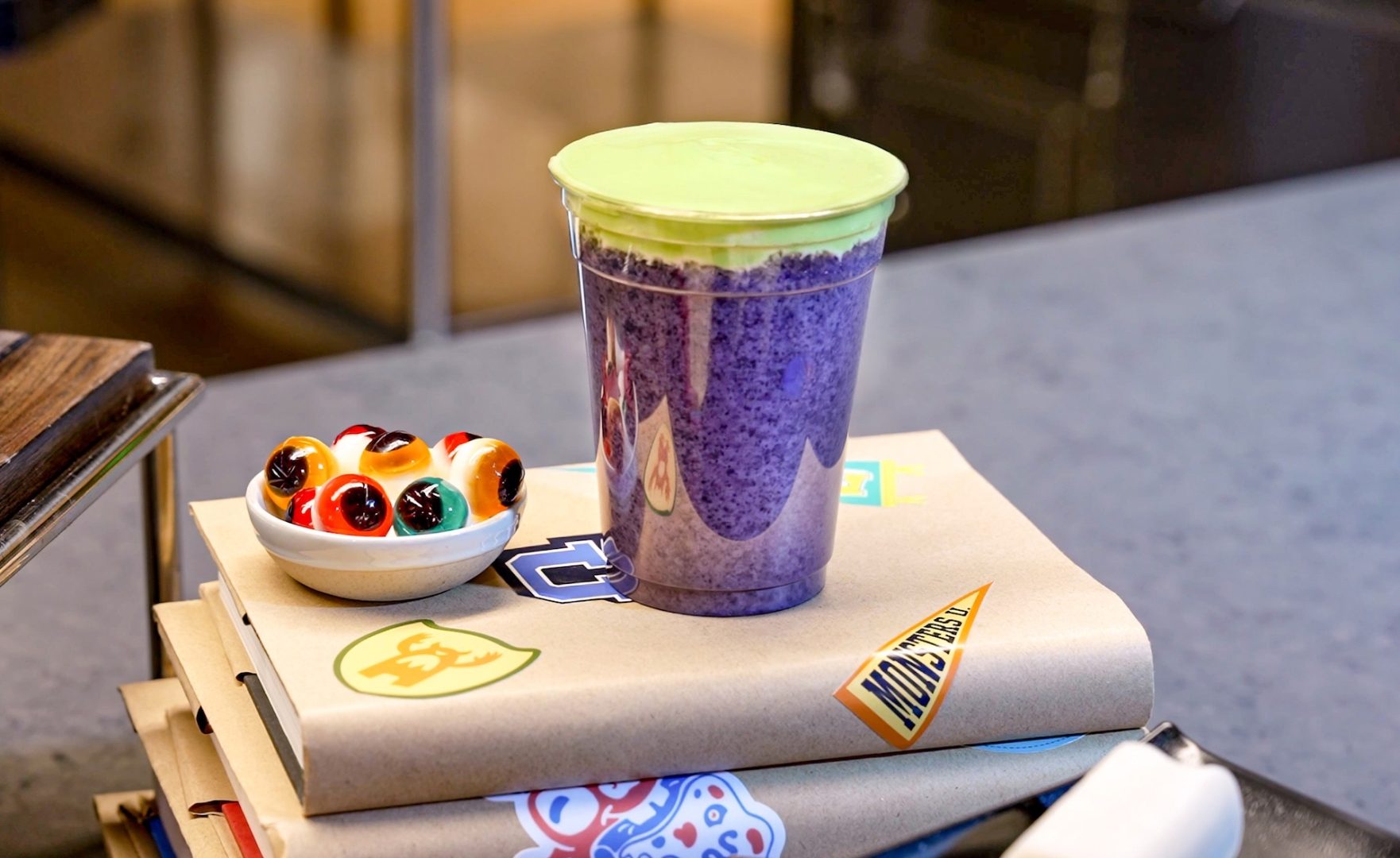 A clear cup of purple frozen drink with a thick lime-green foam topping, on a stack of brown-paper-covered textbooks with Monsters University stickers, next to a small dish of gummy eyeball candies