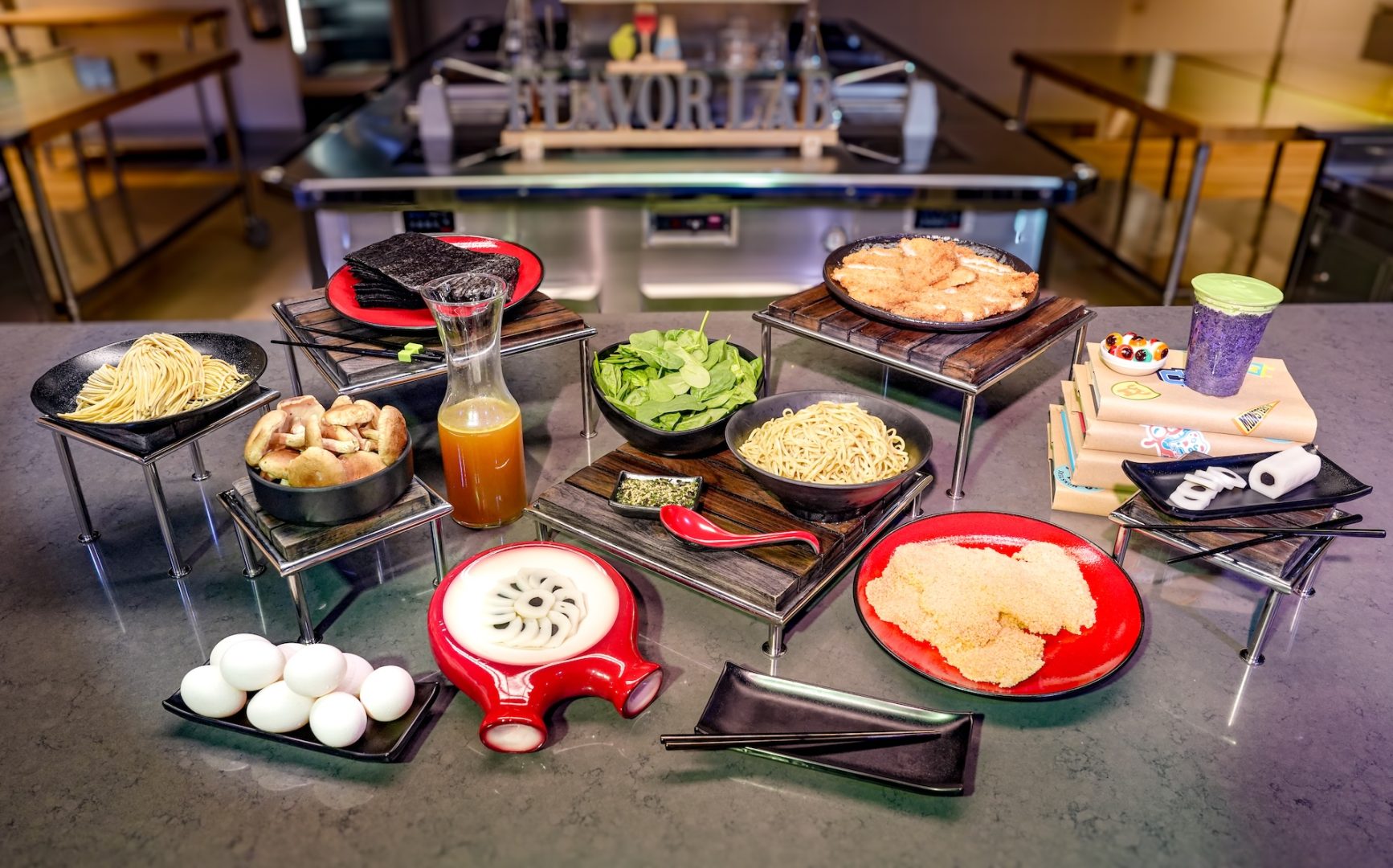 A counter under a Flavor Lab sign, set with ramen noodles, shiitake mushrooms, nori sheets, a carafe of golden broth, spinach, whole eggs, breaded cutlets, sliced fishcake on a red plate, and a purple frozen drink with green foam sitting on a stack of Monsters University textbooks