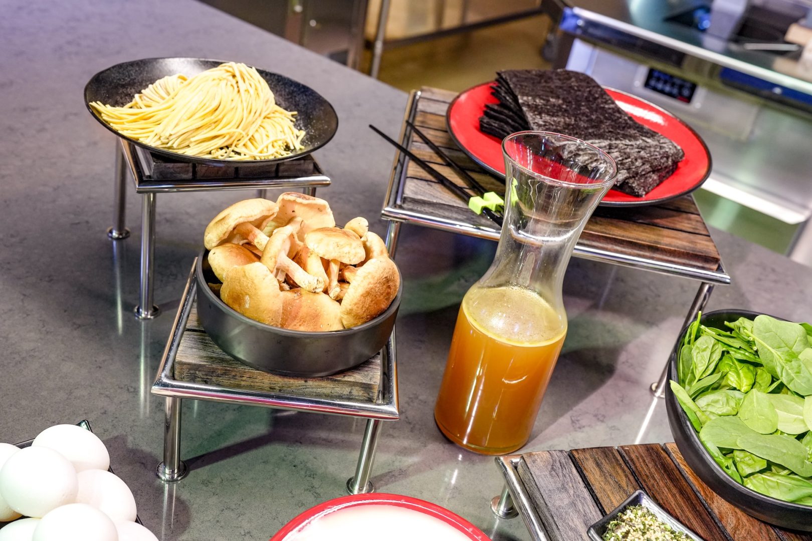 Close-up of fresh ramen noodles on a black plate, a bowl of shiitake mushrooms, a glass carafe of golden broth, a stack of nori sheets on a red plate, and a bowl of spinach on a gray stone counter