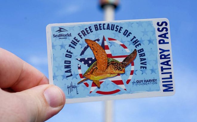 A hand holds up a SeaWorld Orlando Military Pass card showing a sea turtle over a red, white and blue star, with the words Land of the Free Because of the Brave and the Guy Harvey Foundation logo