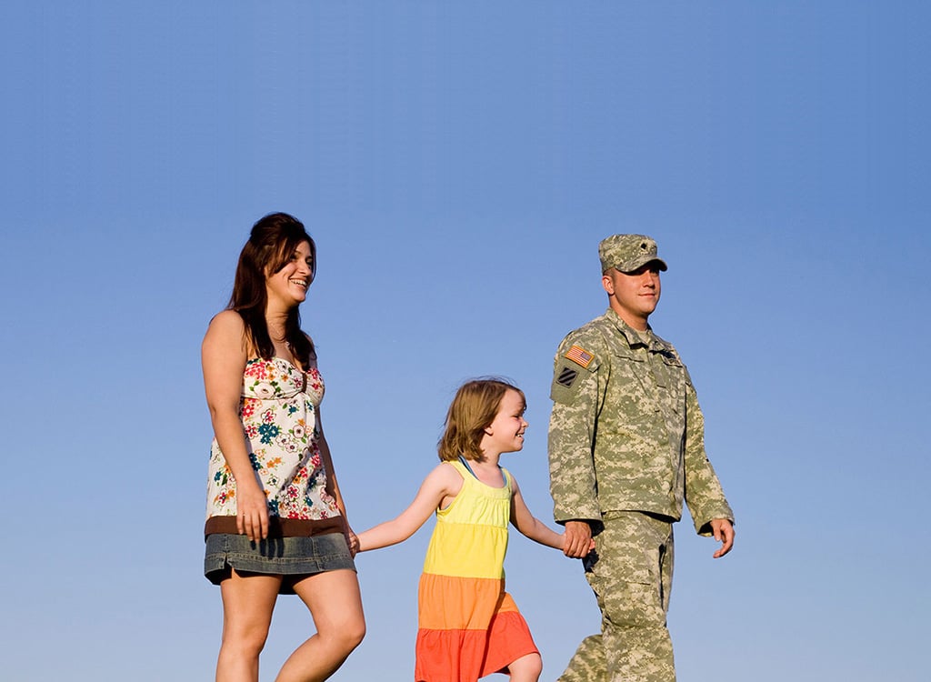 A smiling woman in a floral top and a soldier in camouflage uniform and cap walk with a young girl in a yellow and orange dress between them, holding her hands, against a clear blue sky
