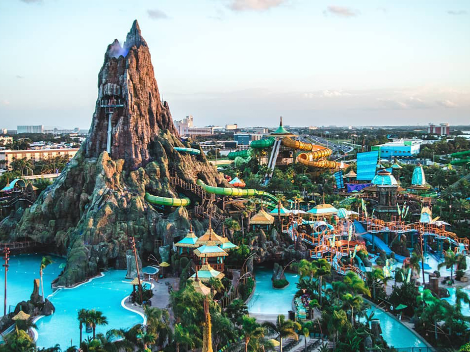 Elevated view of Universal's Volcano Bay in warm low light, with the Krakatau volcano towering over turquoise pools, green and orange water slides and thatched huts, and city buildings on the horizon
