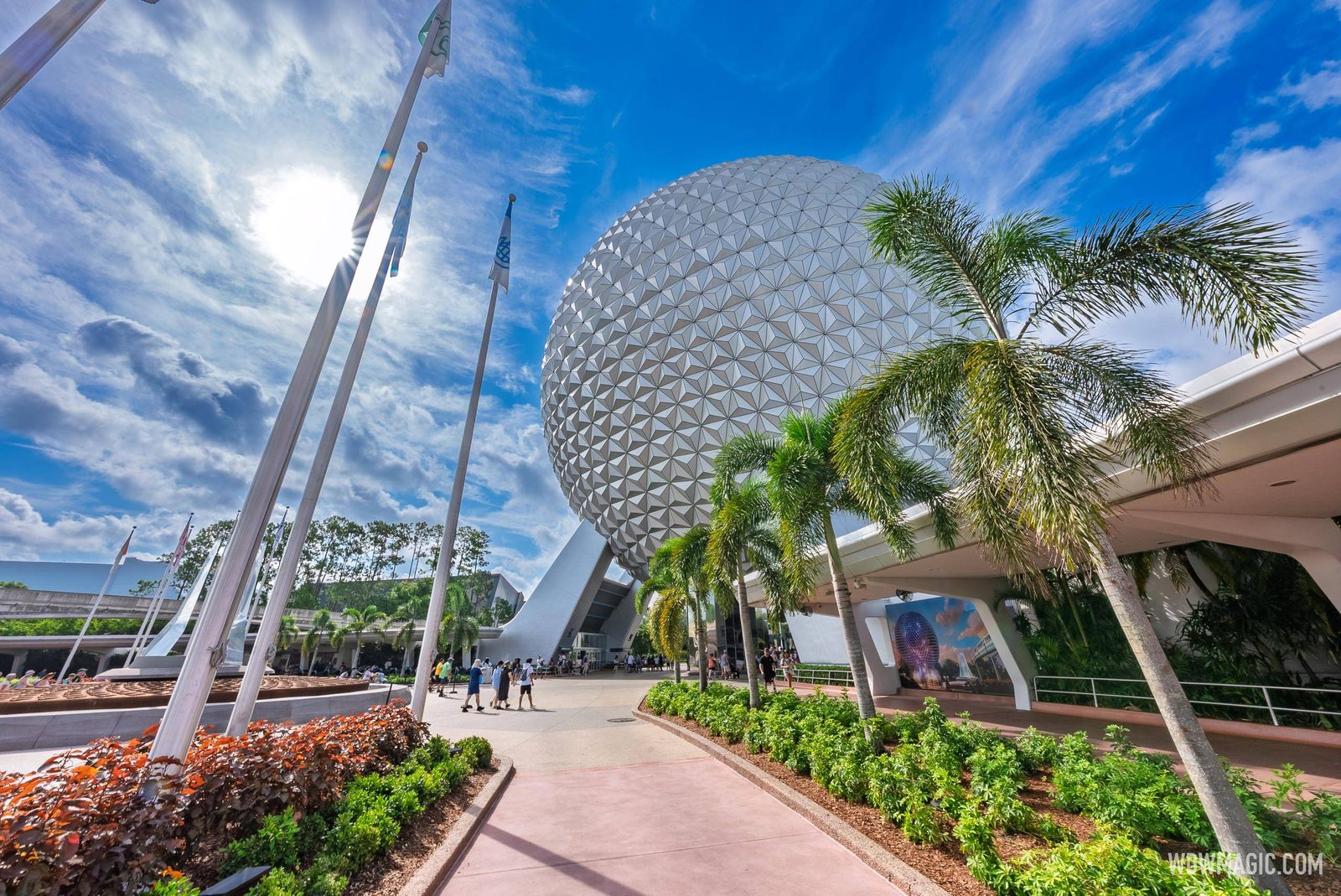 Spaceship Earth rising above palm trees and flagpoles at the EPCOT entrance under a bright blue sky