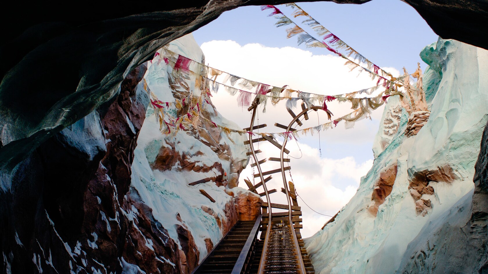 View from a coaster track looking up through a rocky snow-capped mountain opening at a twisted, broken section of track against a blue sky strung with prayer flags
