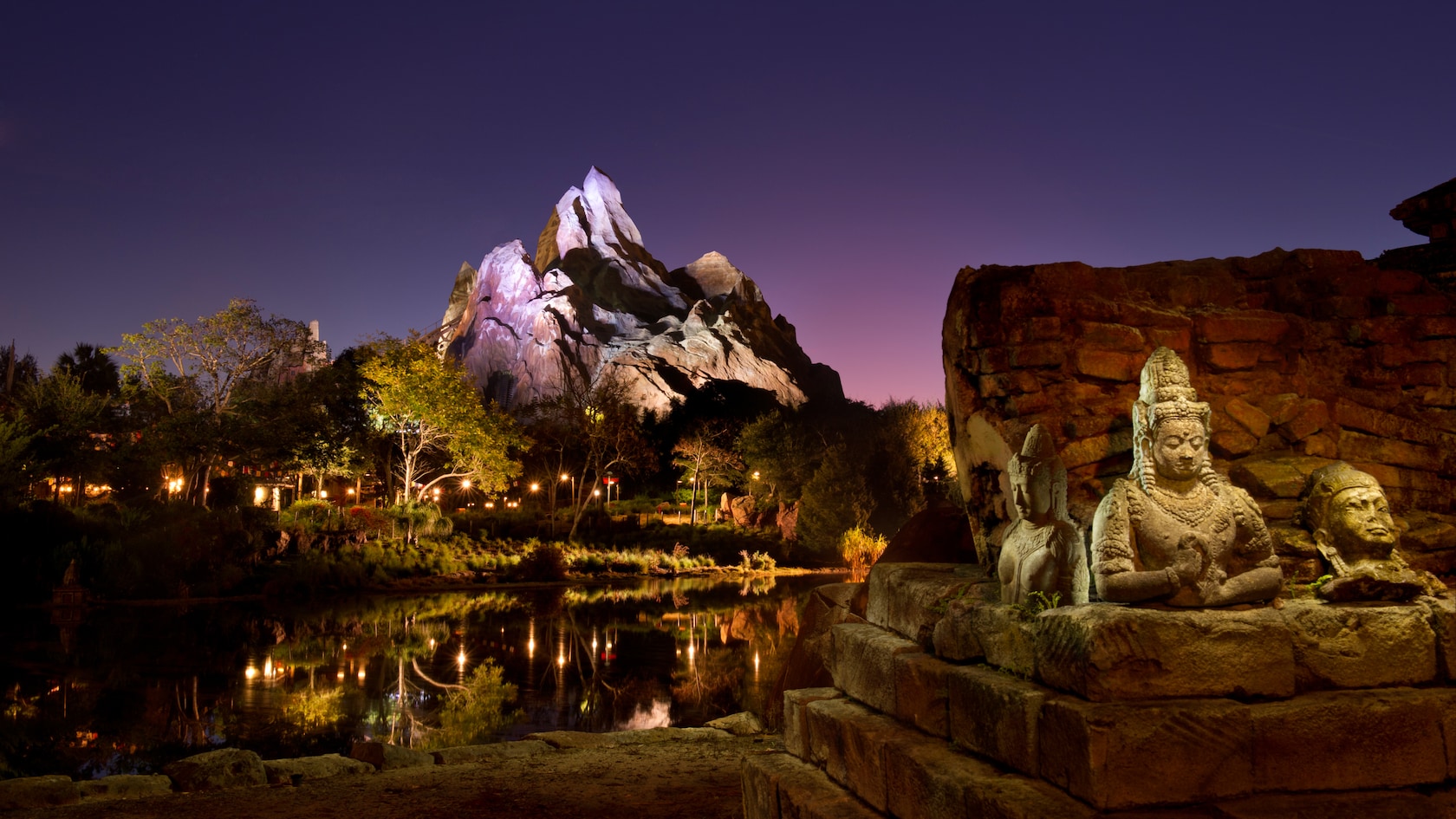 The snow-capped Forbidden Mountain of Expedition Everest lit pale lavender under a purple and pink twilight sky, rising behind trees and still water, with carved stone statues on a ruined wall in the foreground