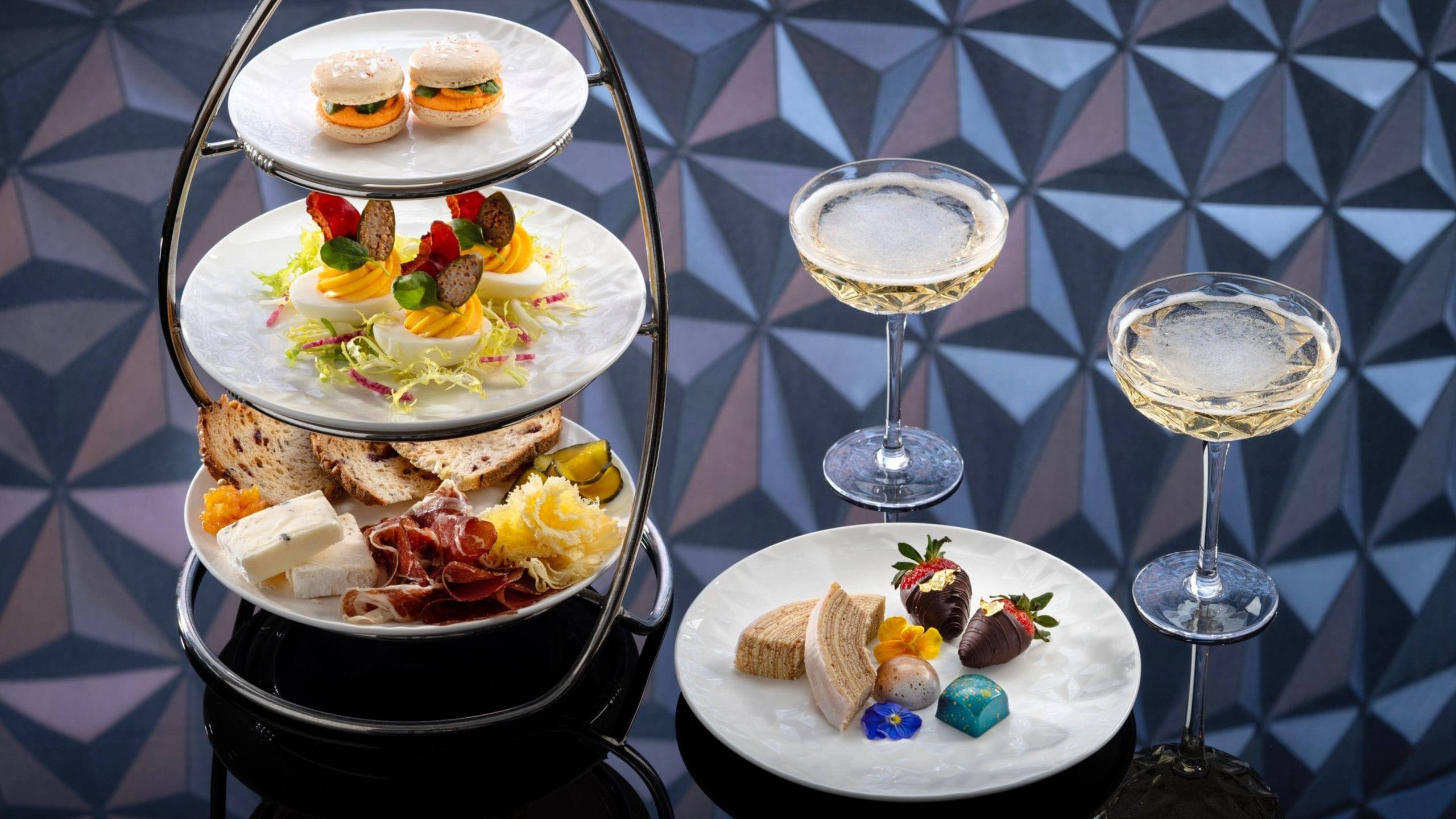 A three-tier tasting tower of savory bites beside a plate of chocolate-dipped strawberries and sweets and two champagne coupes, against a faceted blue and mauve background