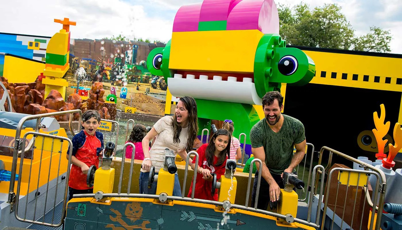 A family laughing as they aim mounted water cannons from a boat on Battle of Bricksburg, with a giant pink, yellow and green brick-built alien figure with big eyes and a toothy grin behind them