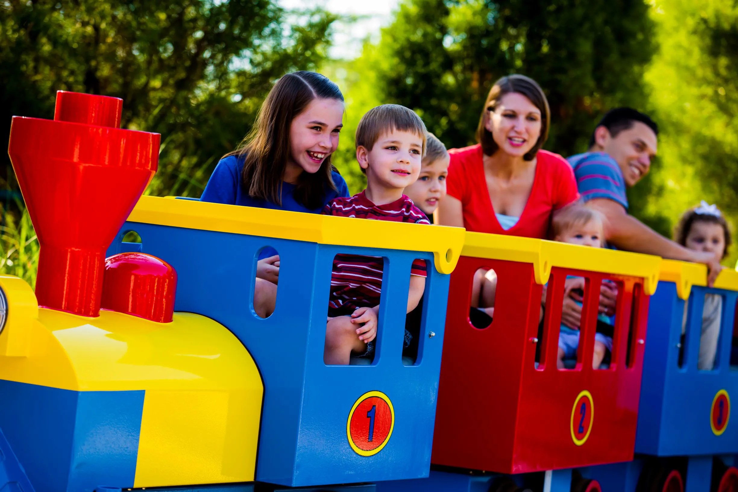 A smiling family riding a toddler train with a yellow and blue engine, a red smokestack and blue and red cars, in front of green trees