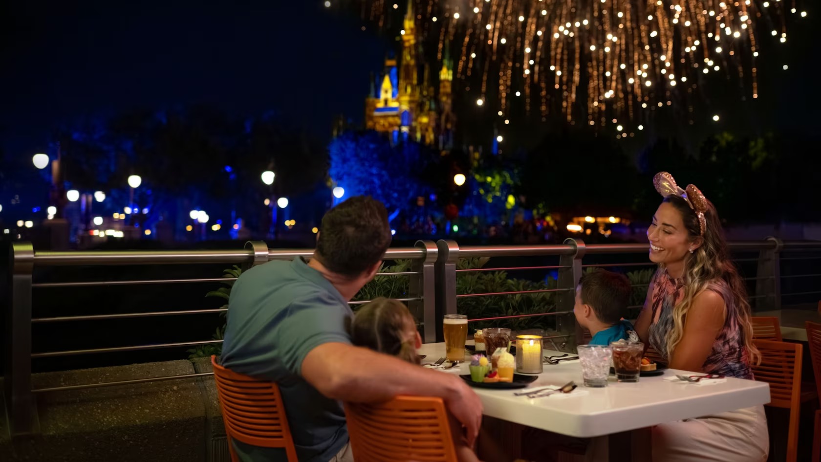 A family at an outdoor table with desserts, drinks and a candle, watching fireworks burst over a lit-up Cinderella Castle at night
