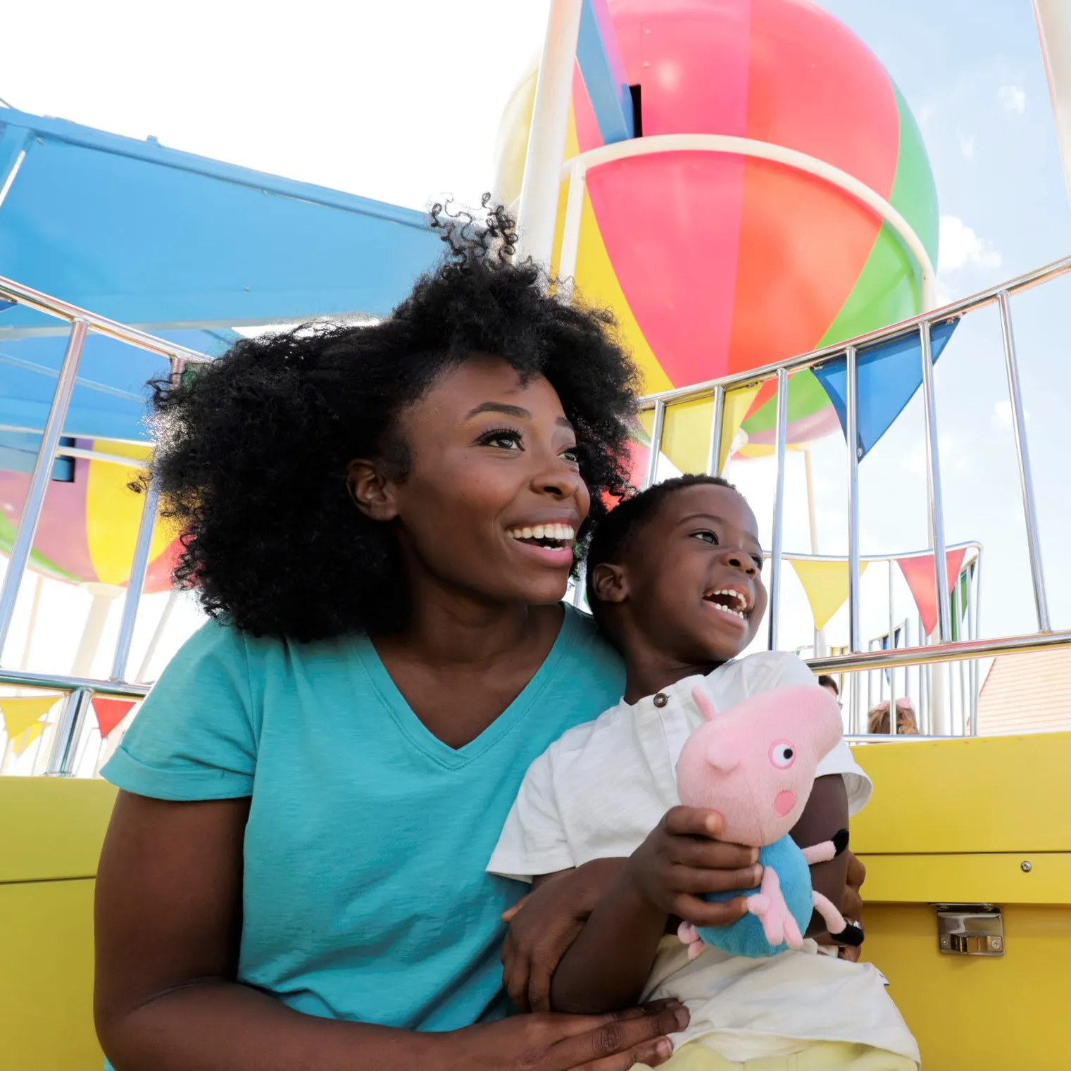 A smiling woman and a laughing young boy holding a George Pig plush toy sit in a yellow ride basket, with a multicolored hot air balloon and bunting behind them