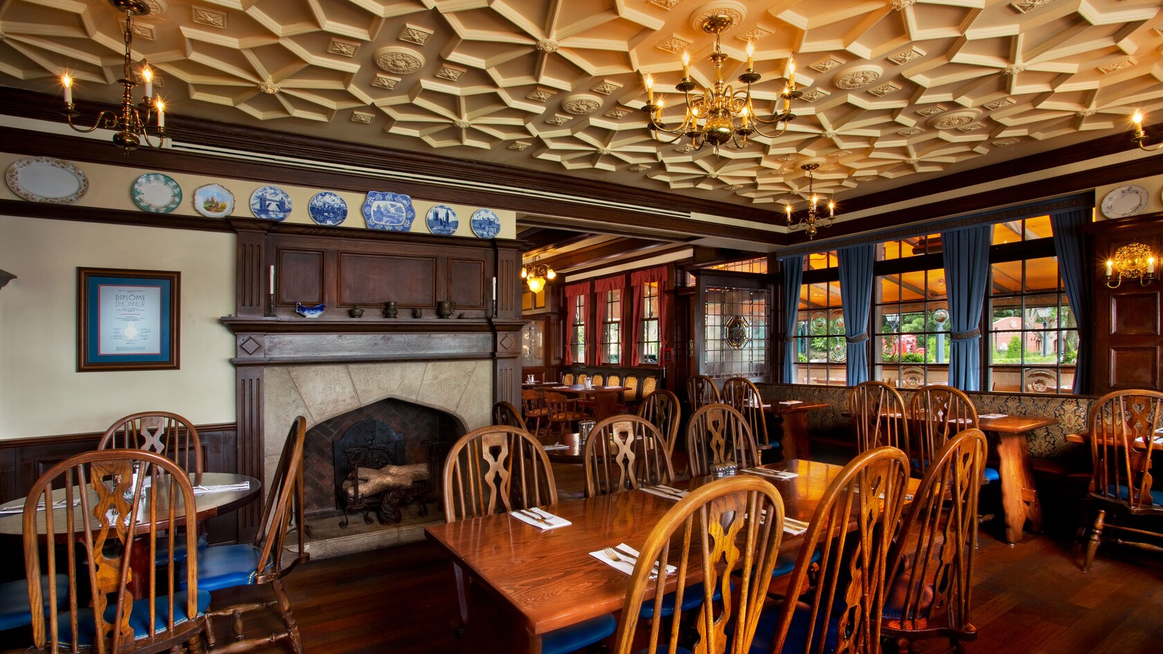 Rose and Crown Dining Room interior with a stone fireplace under a wood mantel, decorative plates on a shelf, brass chandeliers, an ornate geometric ceiling and wooden spindle-back chairs at set tables