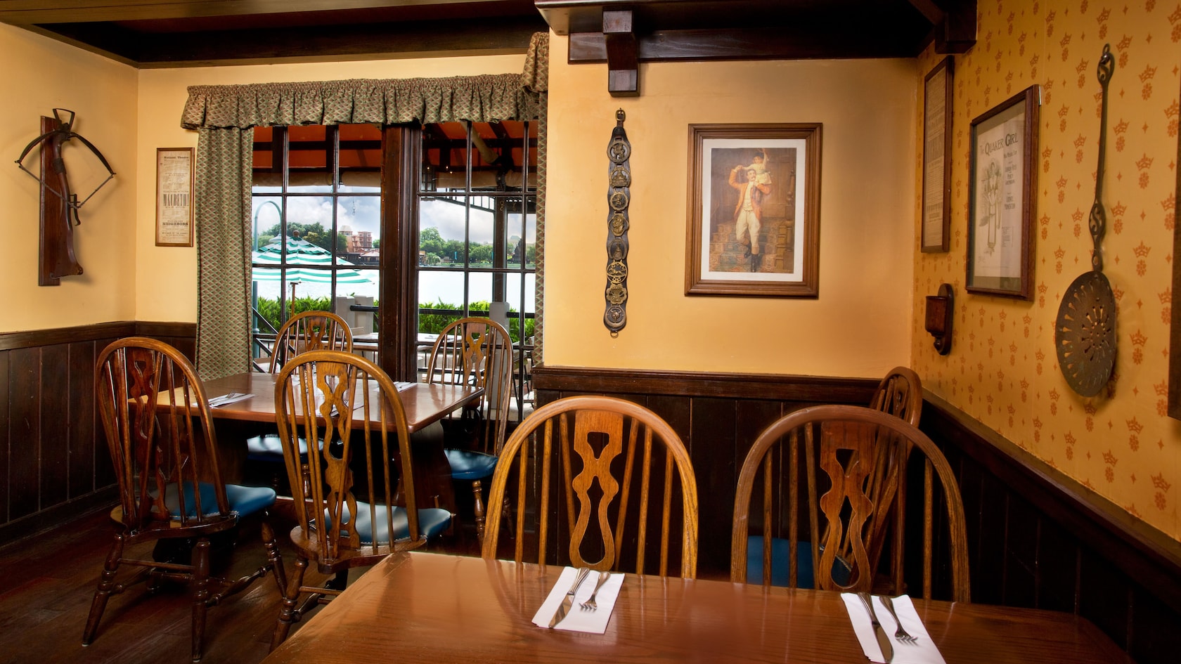 A corner of the dining room with yellow crown-patterned wallpaper, framed vintage prints, a hanging brass utensil and wooden chairs at a table beside a window looking out to the patio and lagoon