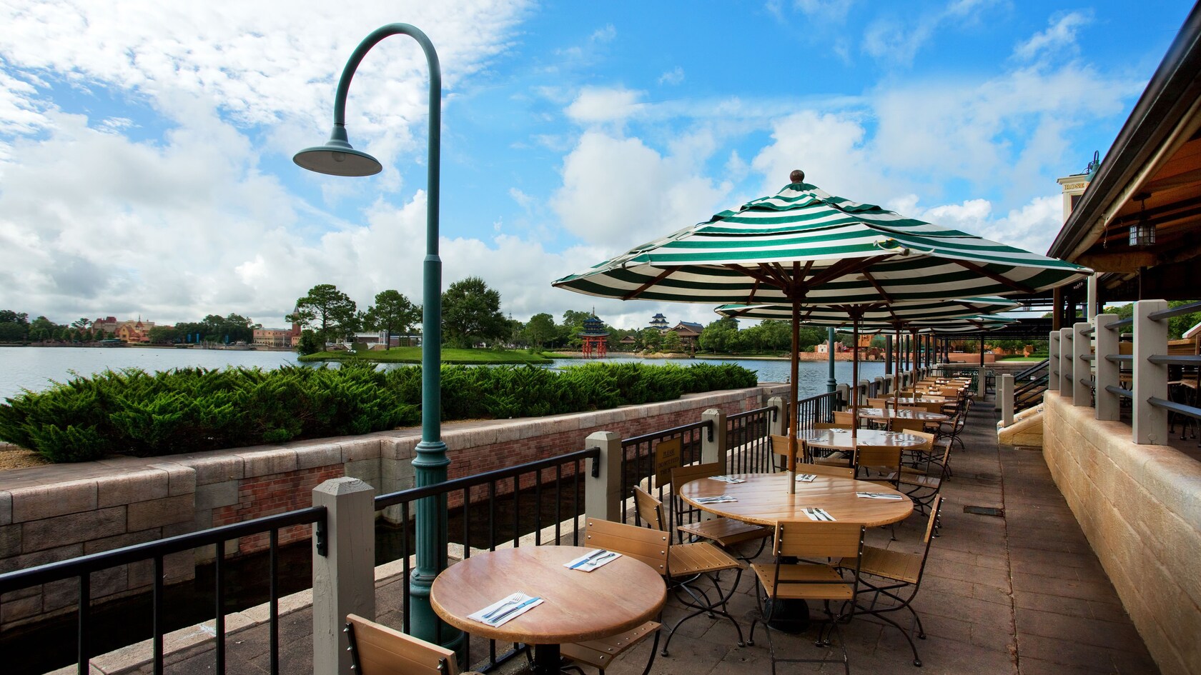 Outdoor patio tables under green and white striped umbrellas beside a railing, with World Showcase Lagoon and the Japan pavilion's red torii gate across the water
