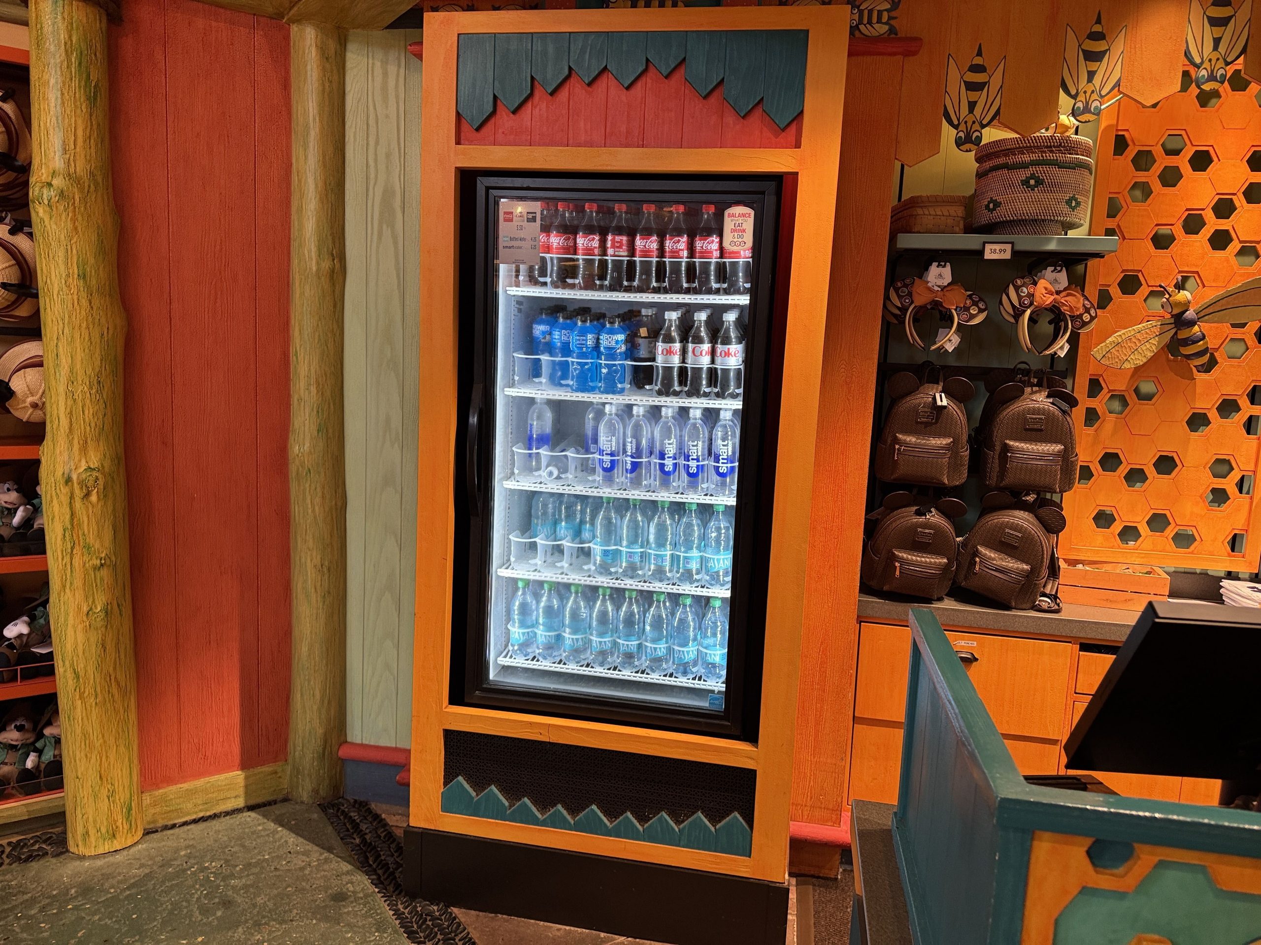 A themed orange and teal drink fridge in an Animal Kingdom gift shop stocked with Coca-Cola, Powerade, Diet Coke, smartwater and Dasani bottles, next to a display of Mickey ear headbands and brown backpacks