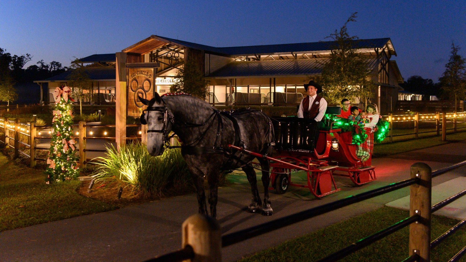 A dapple gray horse pulls a red sleigh trimmed with green lights past a decorated Christmas tree at dusk, with a driver in a cowboy hat, a family riding behind him, and the Tri-Circle-D Ranch barn in the background