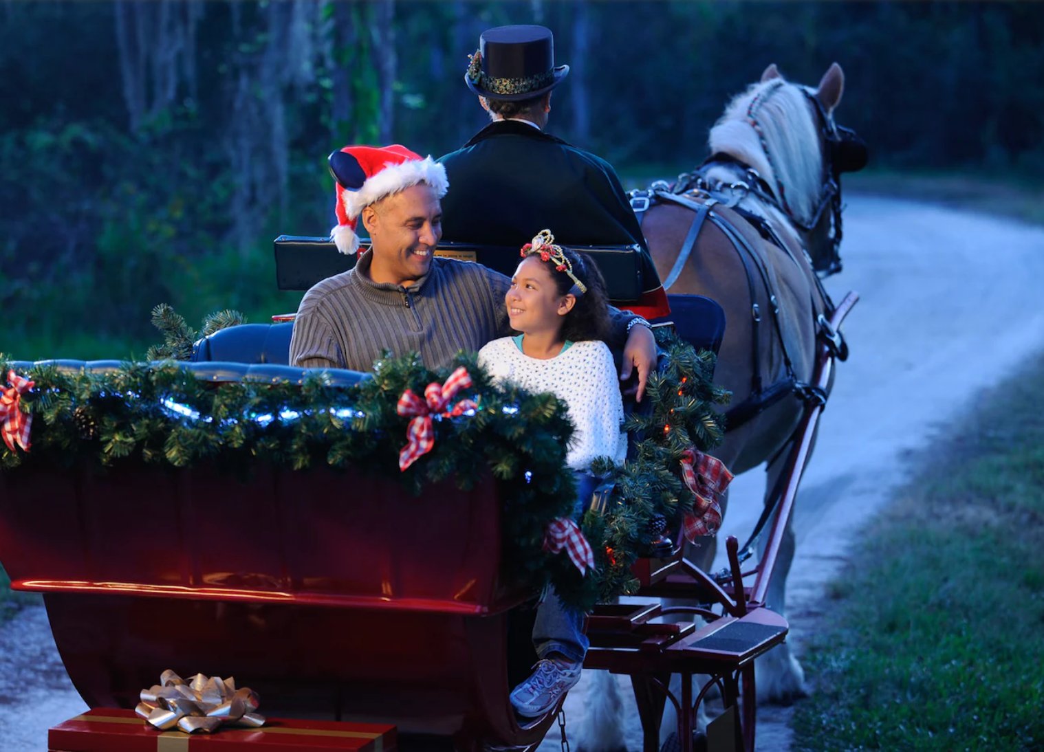 A smiling man in a Santa hat and a girl in a white sweater ride in a red sleigh decorated with garland and plaid bows, with a driver in a top hat and a horse ahead of them on a wooded trail