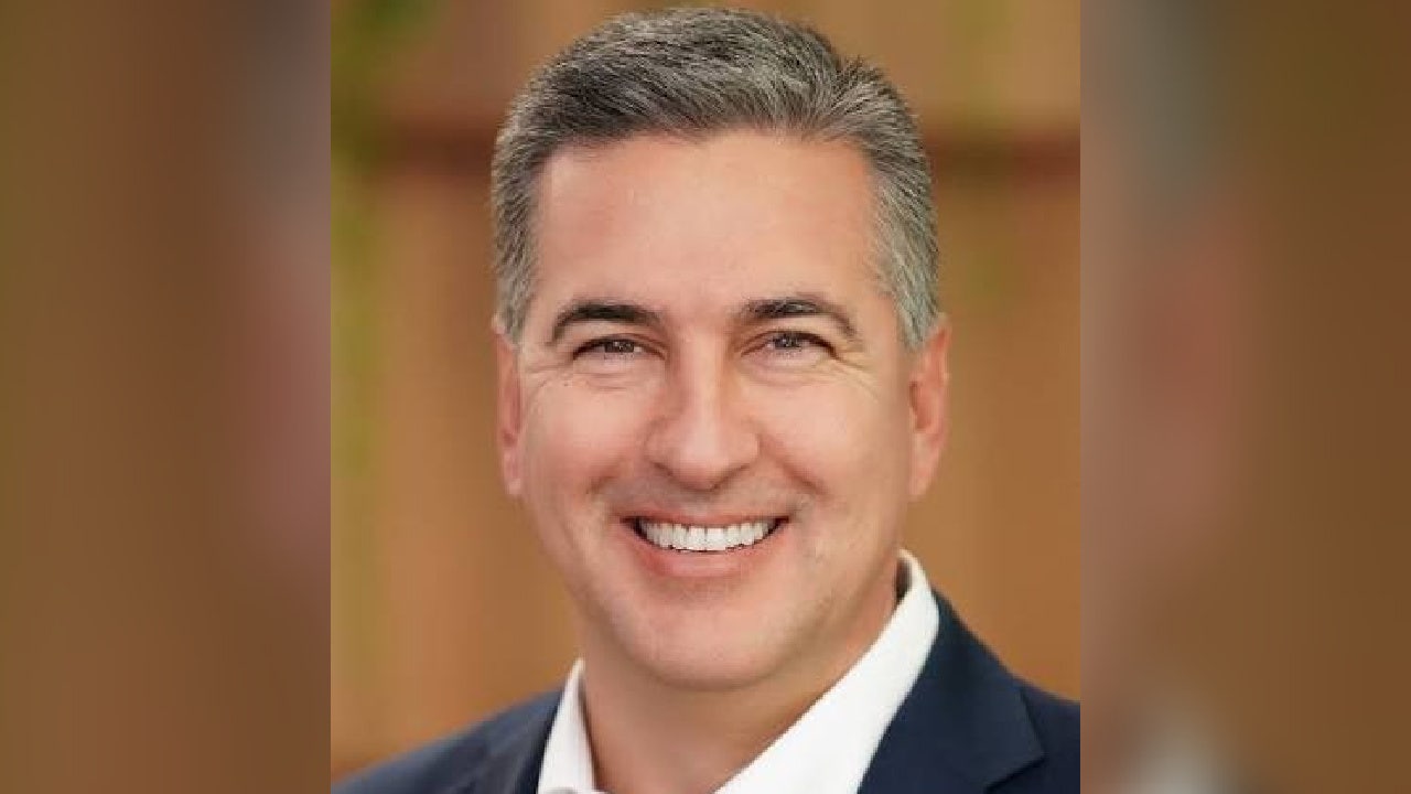 Head-and-shoulders portrait of Brian Bacica, smiling, with short gray hair, a navy jacket and a white open-collar shirt